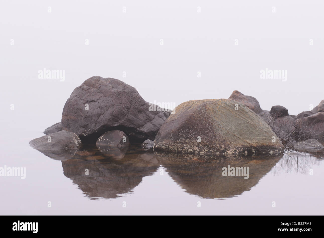 Rock reflecting on a still lake while a thick fog hides the rest of the surroundings. Stock Photo