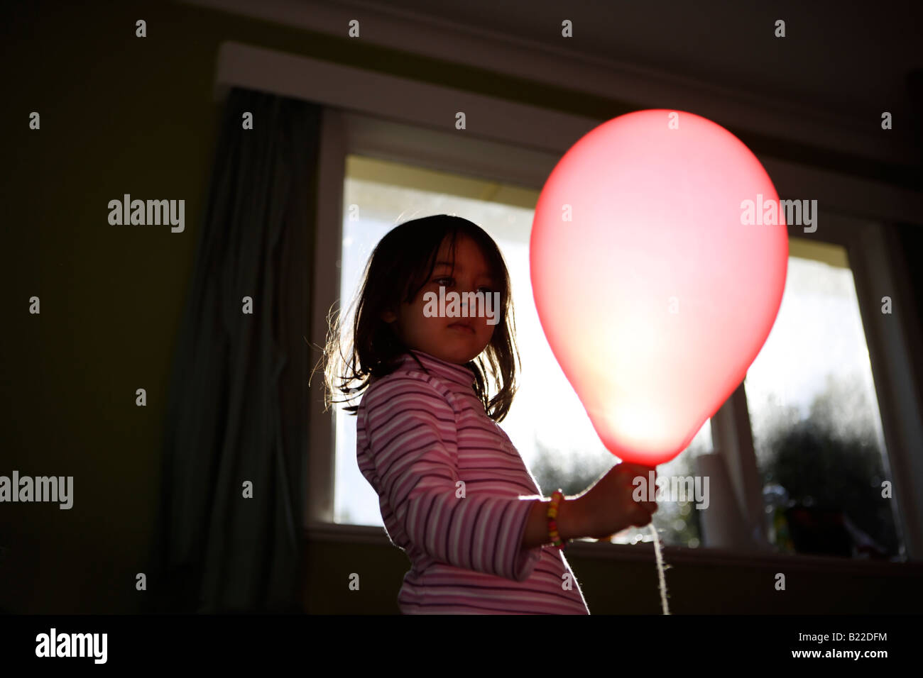 Girl aged four plays with red balloon lit up by afternoon sun streaming through a window. Mixed race asian english Stock Photo