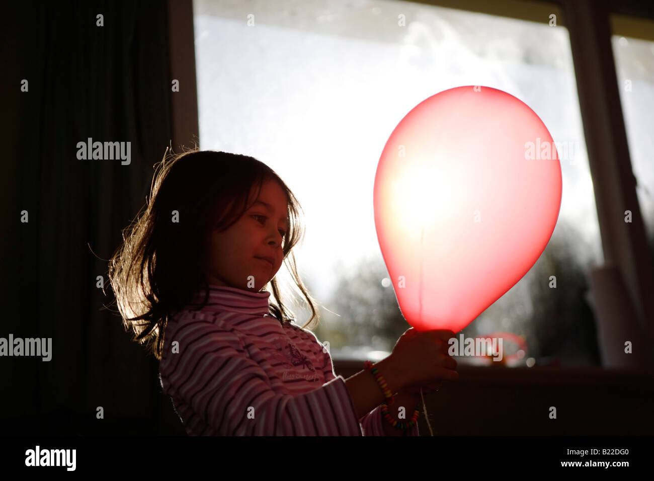 Girl aged four plays with red balloon lit up by afternoon sun streaming through a window Stock Photo