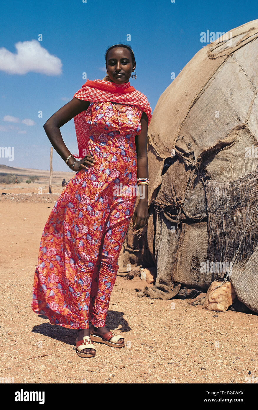Somali woman wears vivid long red patterned dress of modern fabric outside her traditional hut northern Kenya East Africa Stock Photo
