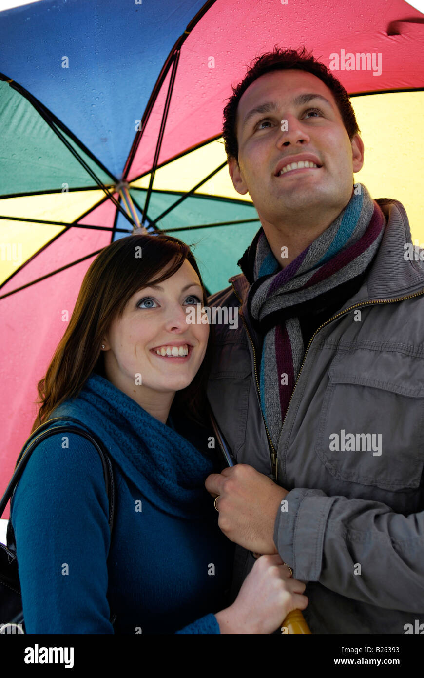 A happy young couple looking up at the sky from under the shelter of a large colourful umbrella Stock Photo
