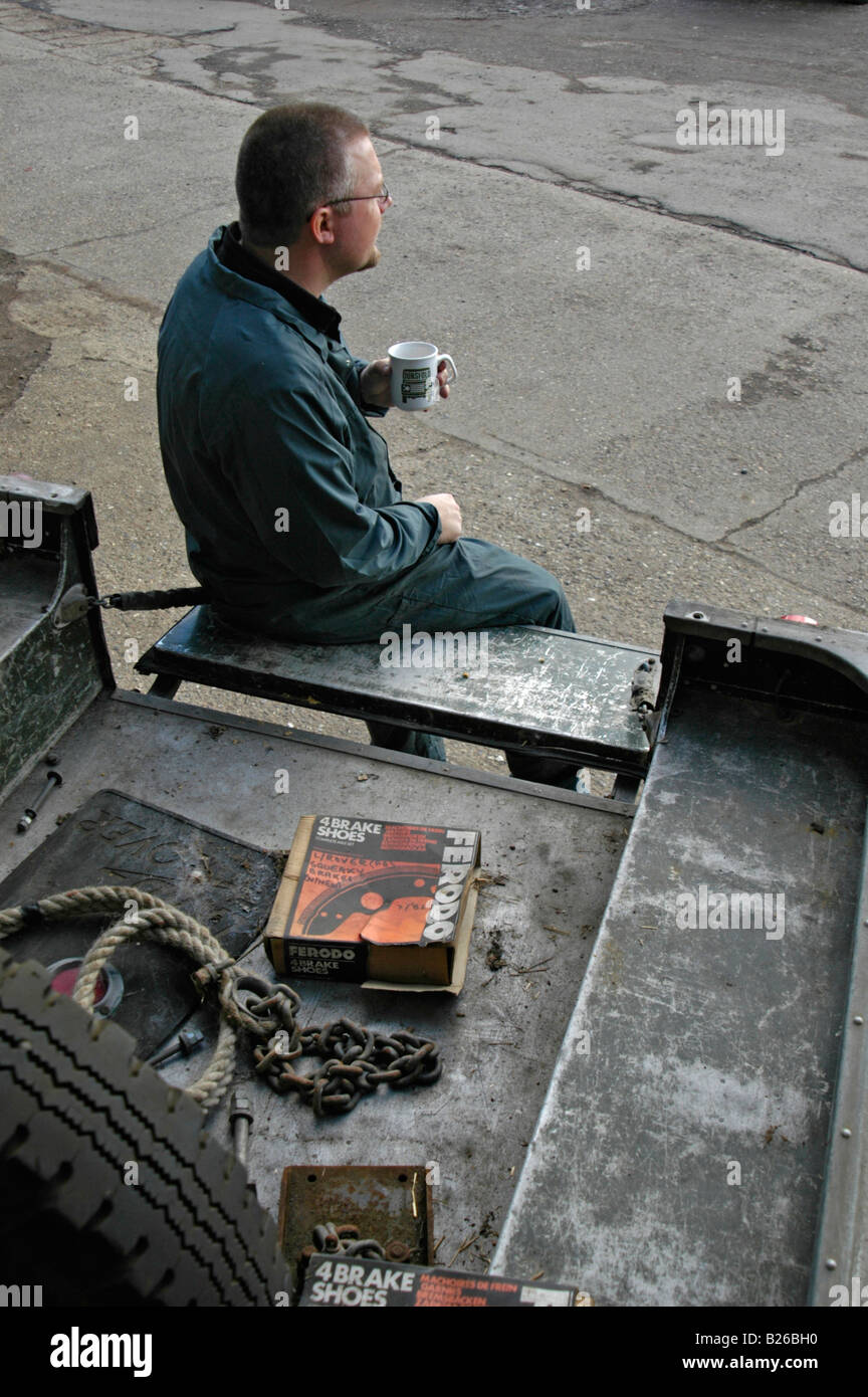 Car mechanic having a morning break on the loadbed of a 1950's Land Rover Series One truck. Stock Photo