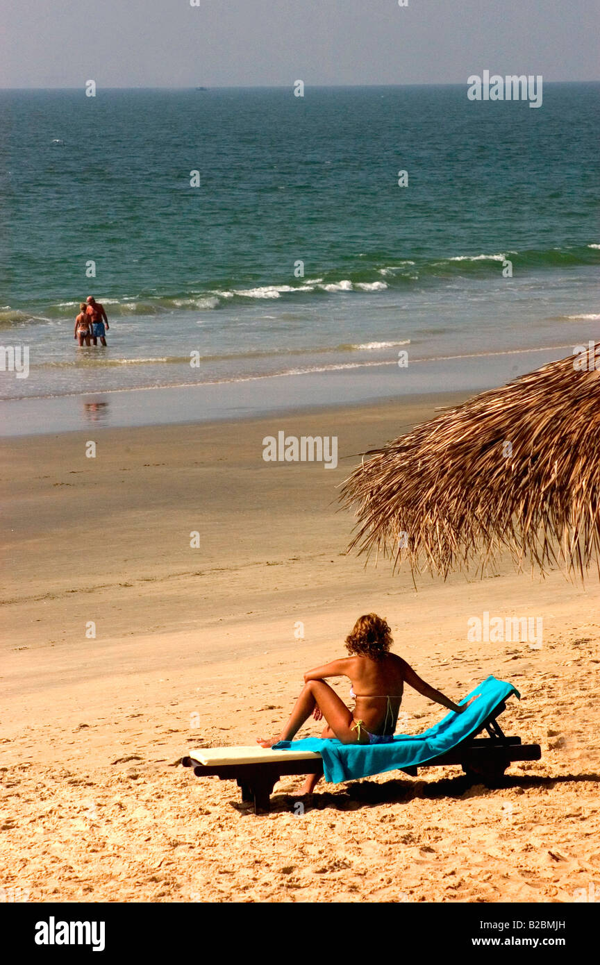Majorda Beach South Goa Woman sitting on sunbed beneath raffita beach umbrella looking out to sea Couple standing in sea Stock Photo