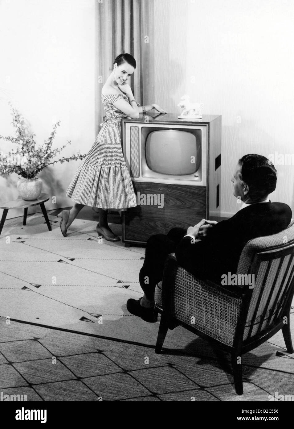 Woman posing in front of a TV set, man sitting in an armchair, historical photo, circa 1955 Stock Photo