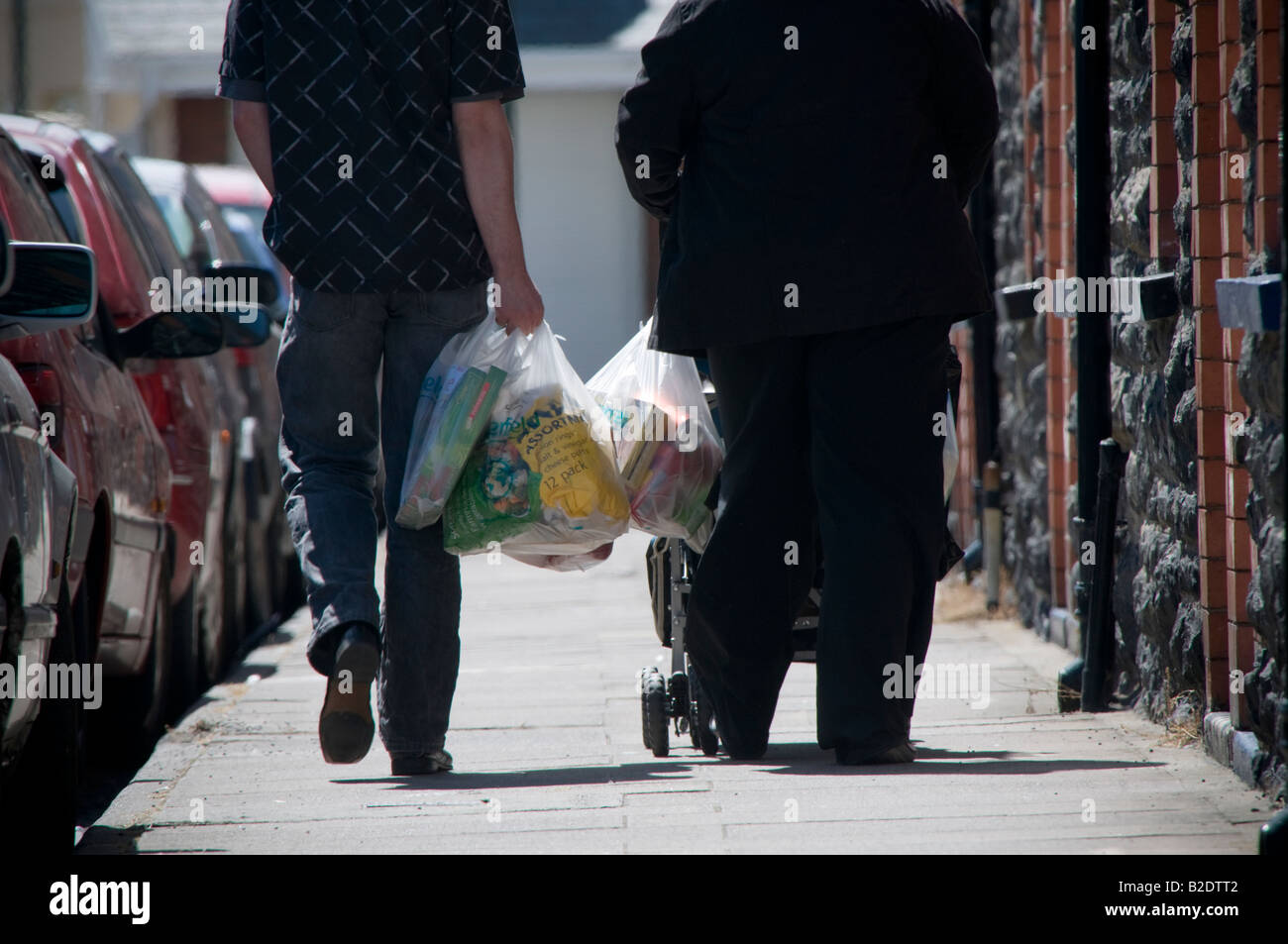 young couple family walking home from supermarket with shopping in plastic bags and pushing a childs buggy rear view, UK Stock Photo