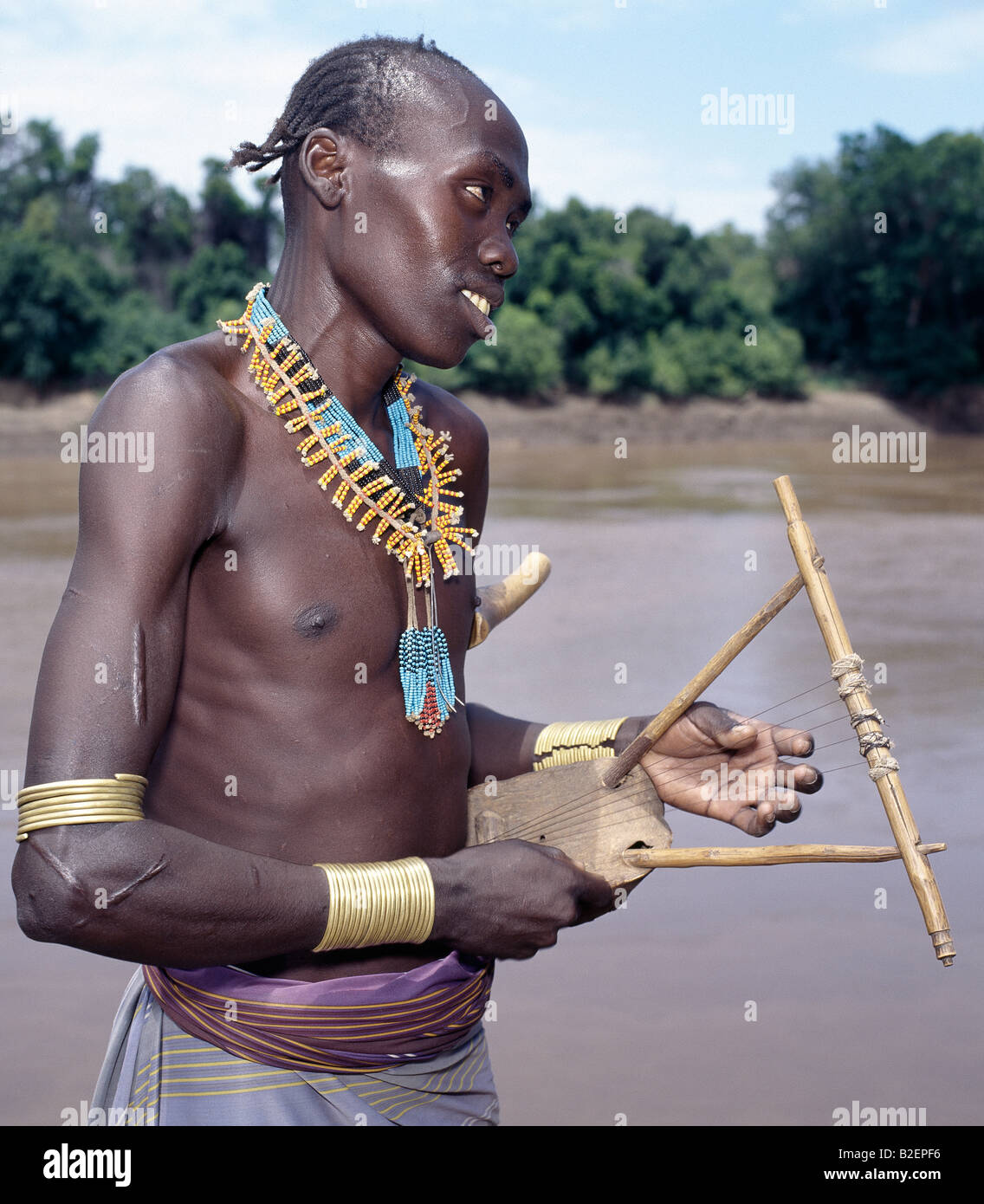 A Karo man with braided hair plays a traditional stringed instrument beside the Omo River near Duss. Stock Photo