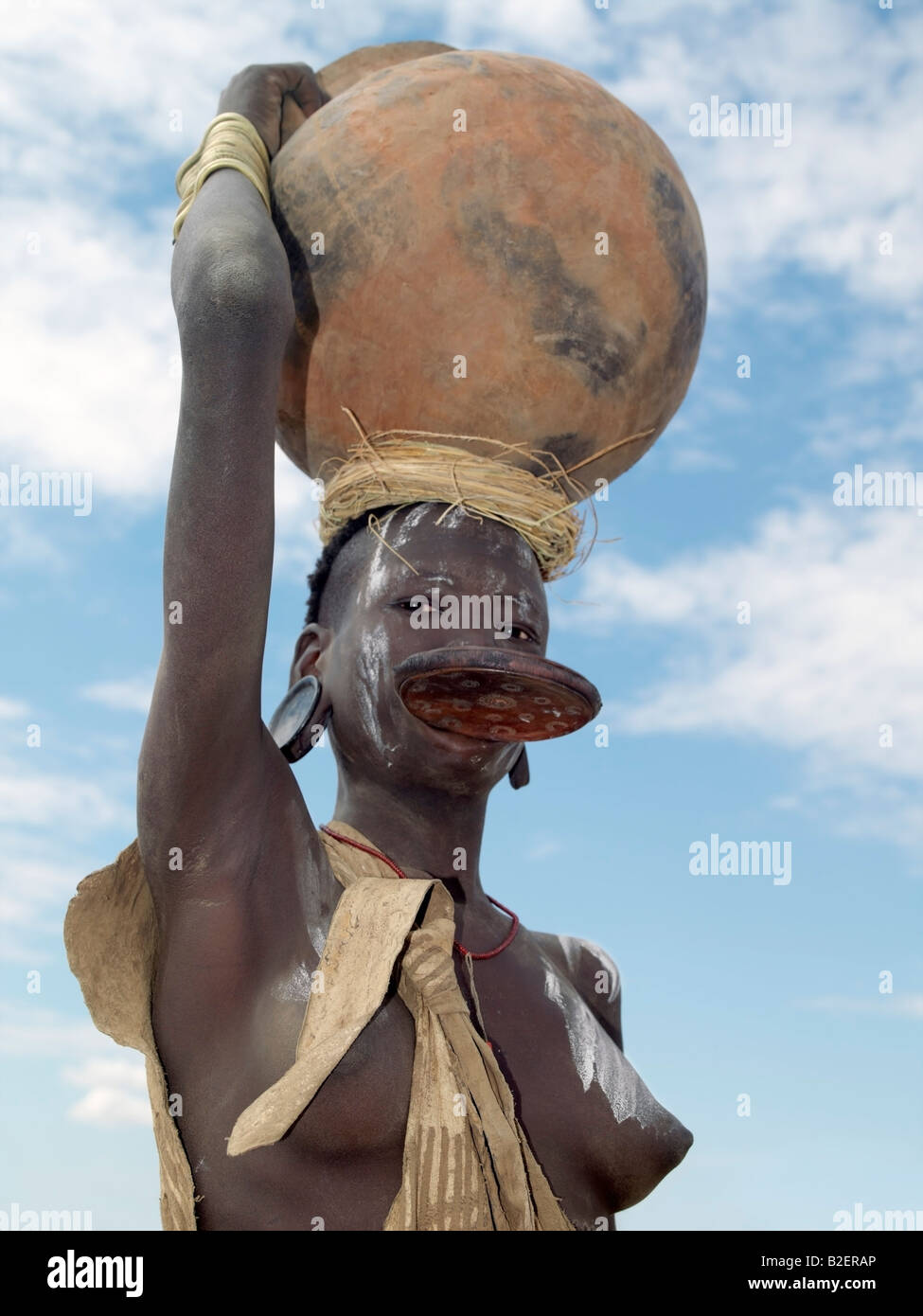 A Mursi woman wearing a large clay lip plate and ear ornaments to match carries a large clay pot on her head. Stock Photo