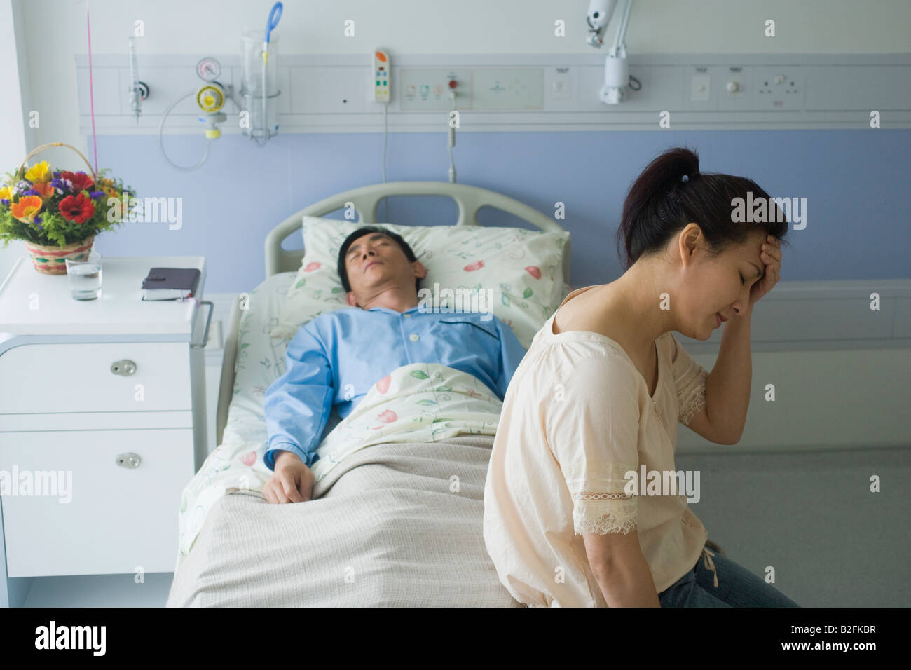 Mid adult man sleeping on the bed and a mid adult woman looking sad in a hospital room Stock Photo