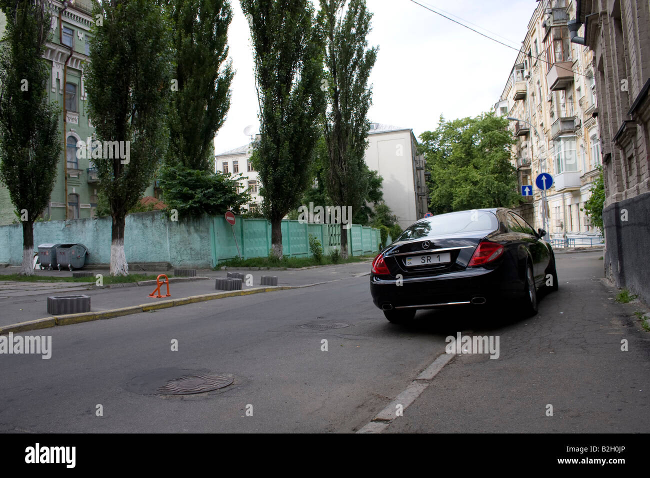 Quiet street in Kyiv with tall trees, residential buildings, a green fence, trash bins, and a black luxury car parked along the sidewalk. Stock Photo