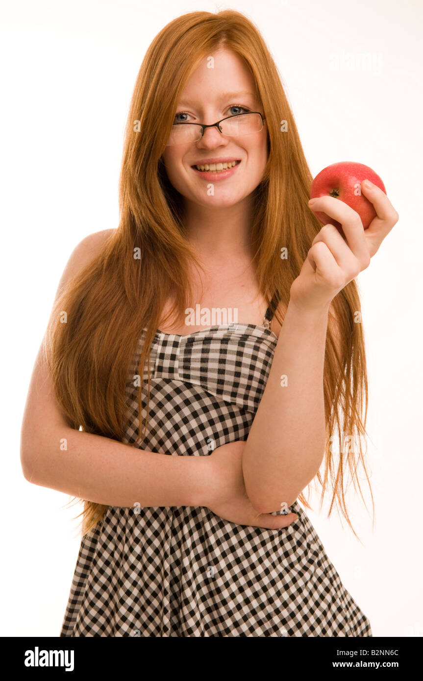 Red haired 17 18 19 year old slim pretty teenage girl holding apple smiling wearing glasses long hair, white background UK Stock Photo