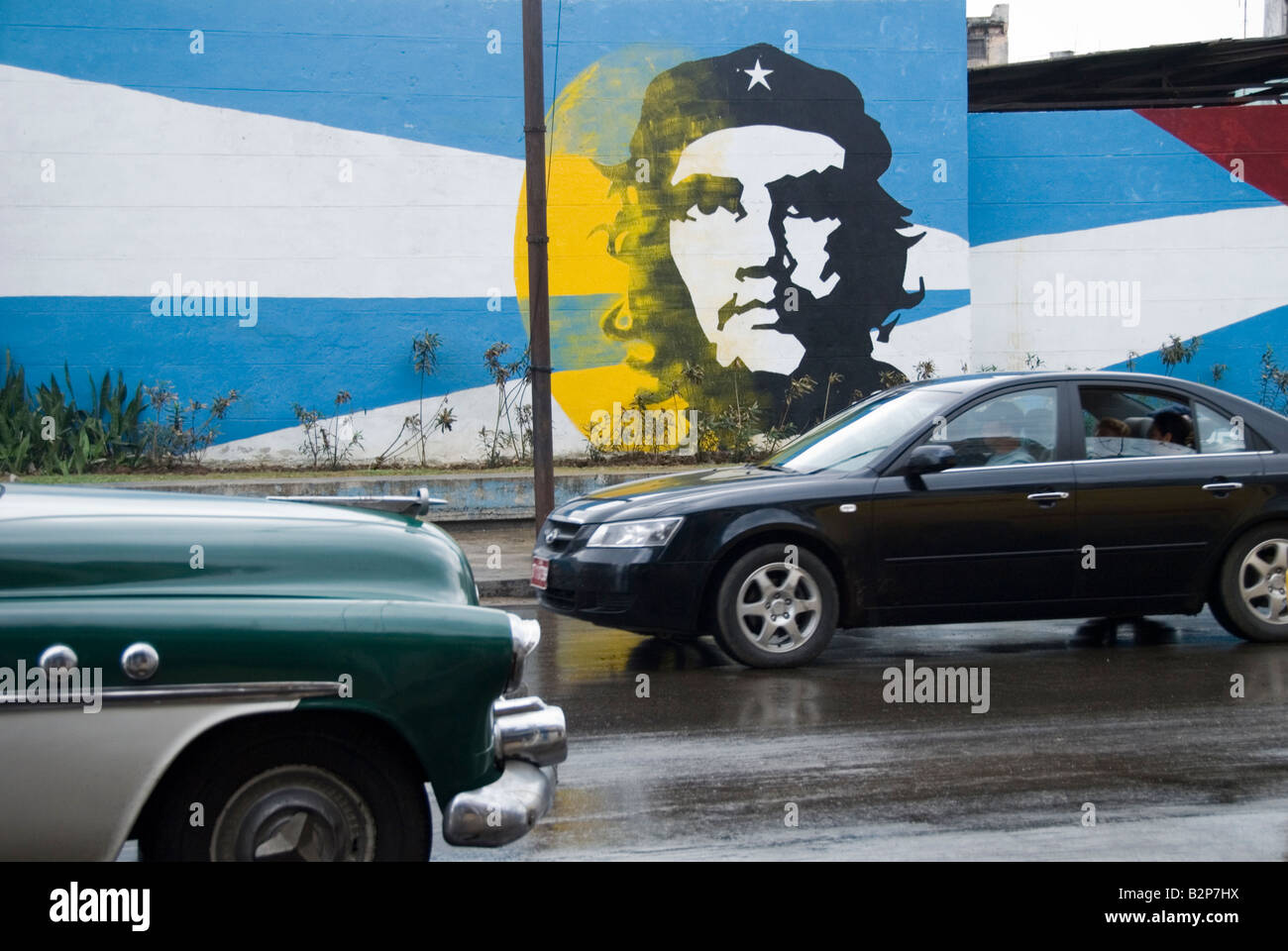Vintage car and new car passing by a mural of Ernesto Che Guevara during time of embargo in Cuba Central Havana Cuba Stock Photo