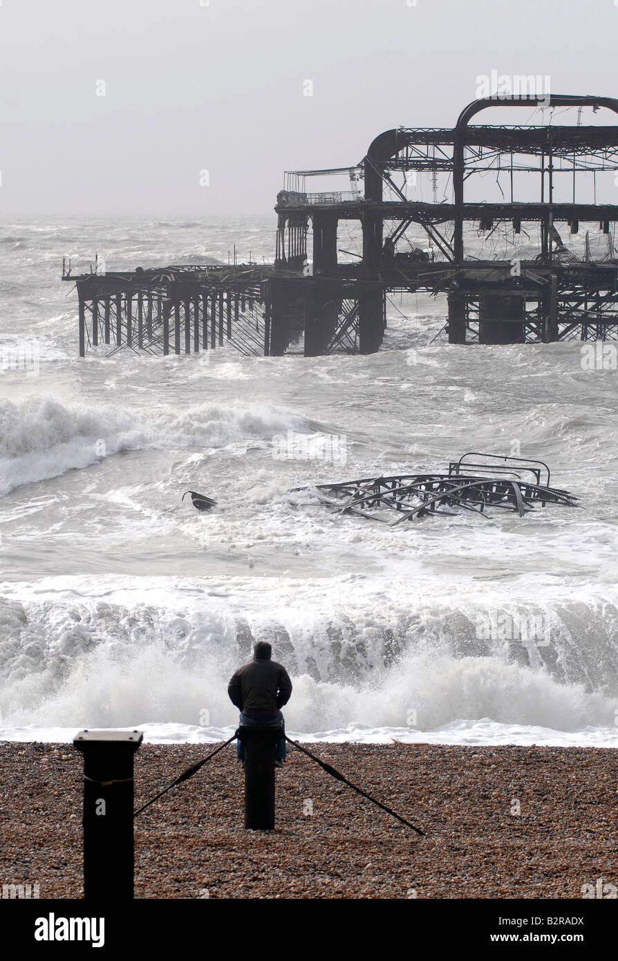 A man watches waves on the beach by the ruins of the West Pier during a winter storm Brighton UK Stock Photo