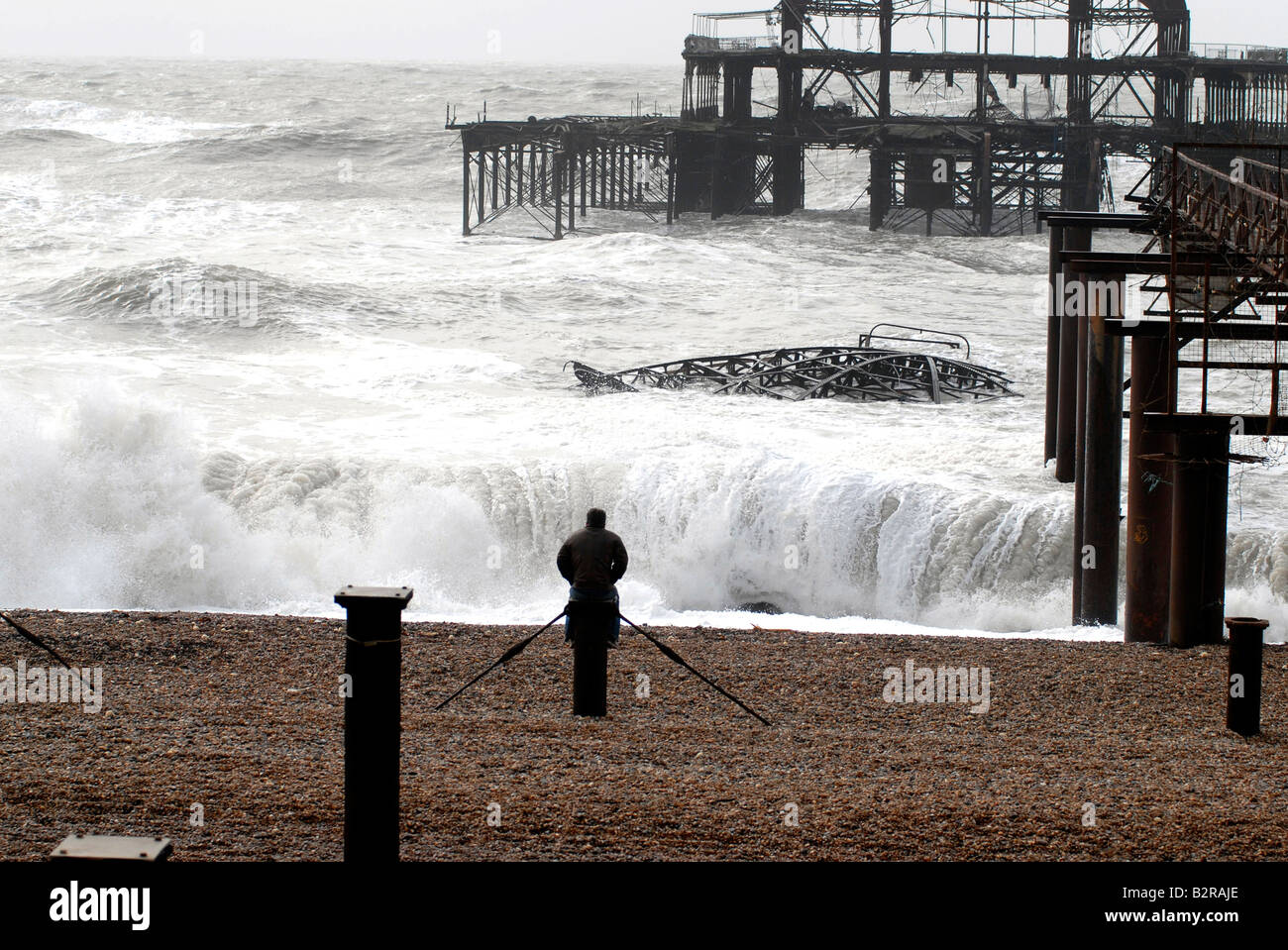 A man watches waves on the beach by the ruins of the West Pier during a winter storm Brighton UK Stock Photo