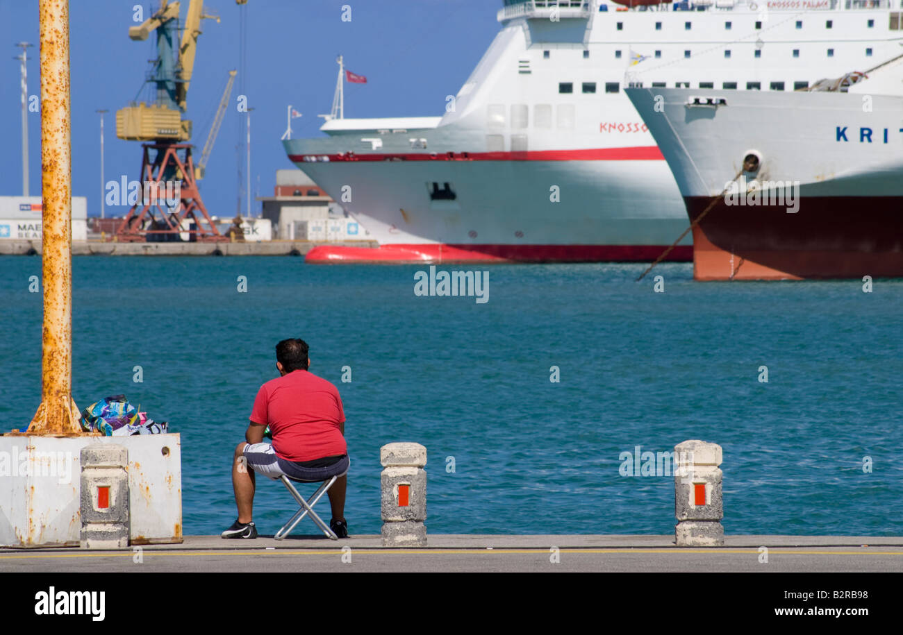 Heraklion, Iraklio, Crete, Greece. Man fishing in harbour; cruise ships in background Stock Photo