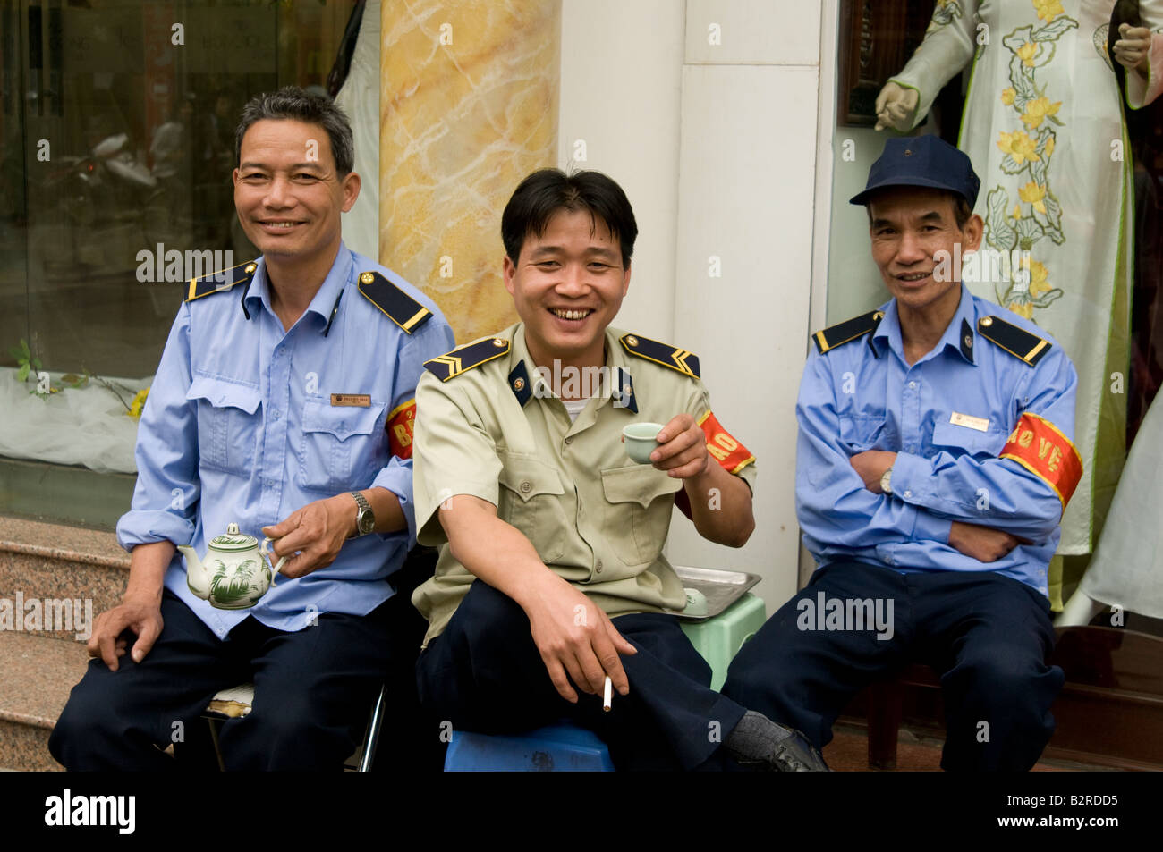 Three Vietnamese security guards sitting in front of a shop drinking tea and laughing on the streets of Hanoi Vietnam Stock Photo