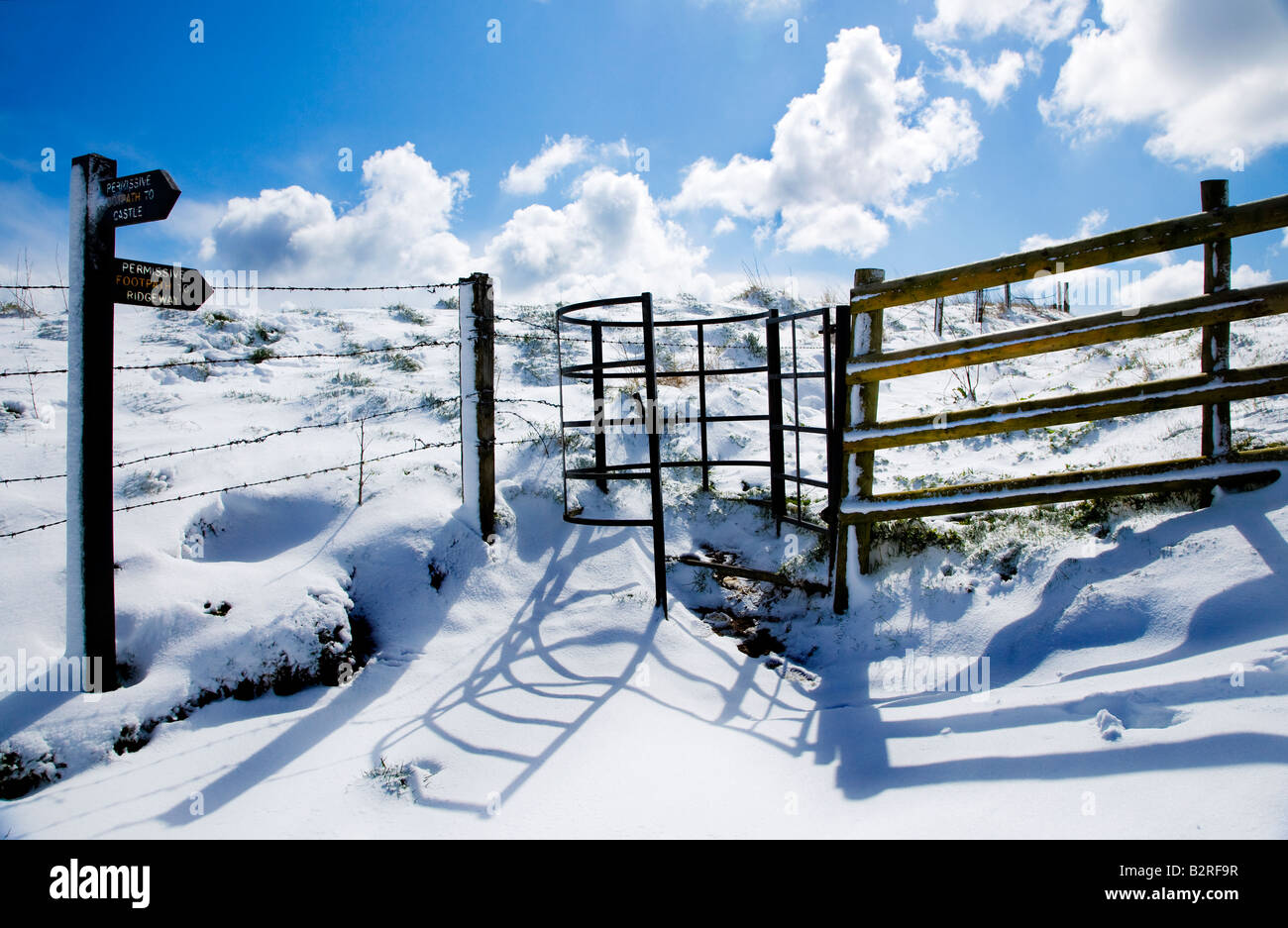 Shadows on newly fallen snow cast by a wrought iron kissing gate and fence with a signpost. Stock Photo