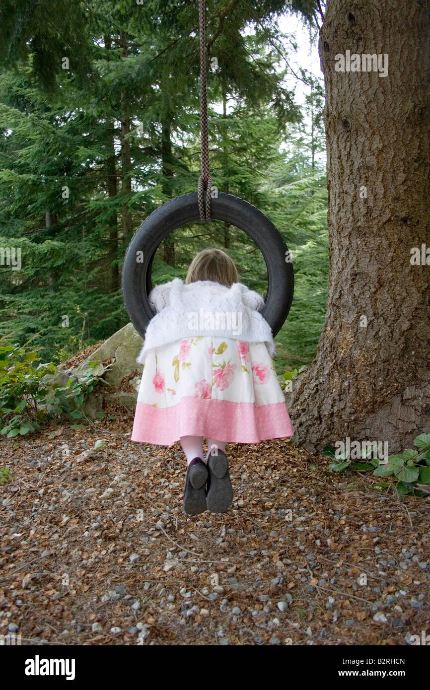young girl wearing pretty dress outside in nature swinging backward on tire swing. child is seen from rear or back view Stock Photo