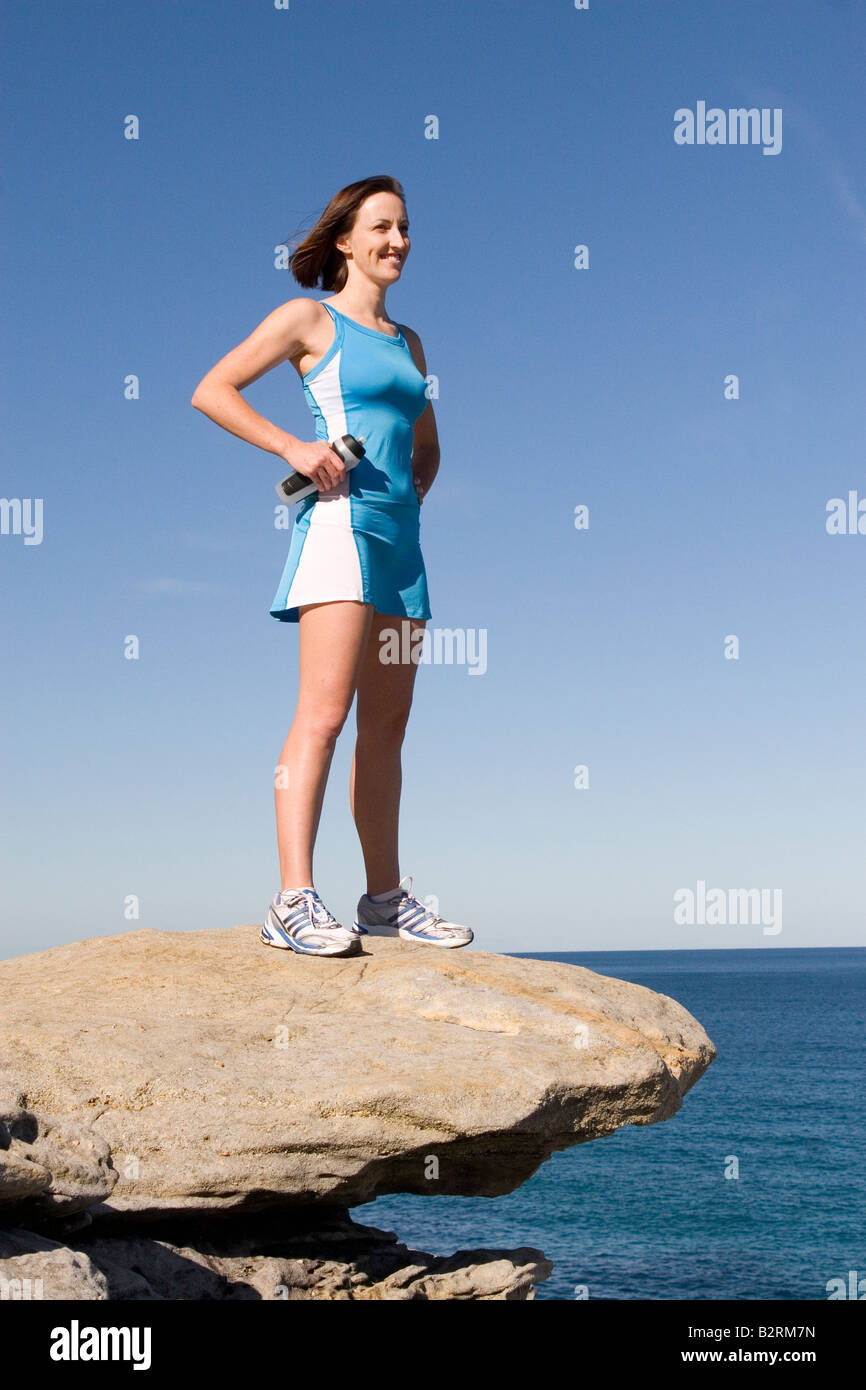 Female Runner in Blue Dress with Water Bottle on Rocky Ledge by Sea Stock Photo