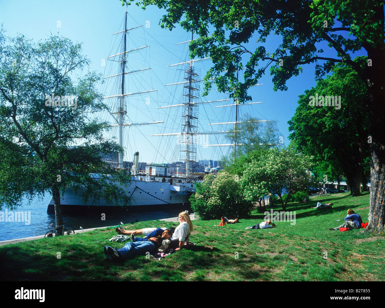 Couples on grass sharing romantic summer moments next to Af Chapman schooner moored at Skeppsholmen Island in Stockholm Stock Photo