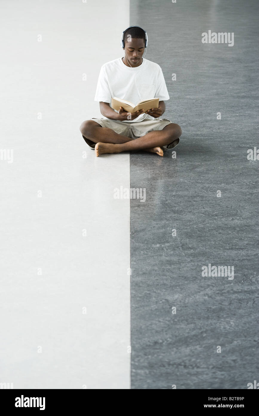 Teenage boy sitting cross-legged on the floor, reading a book, listening to headphones Stock Photo