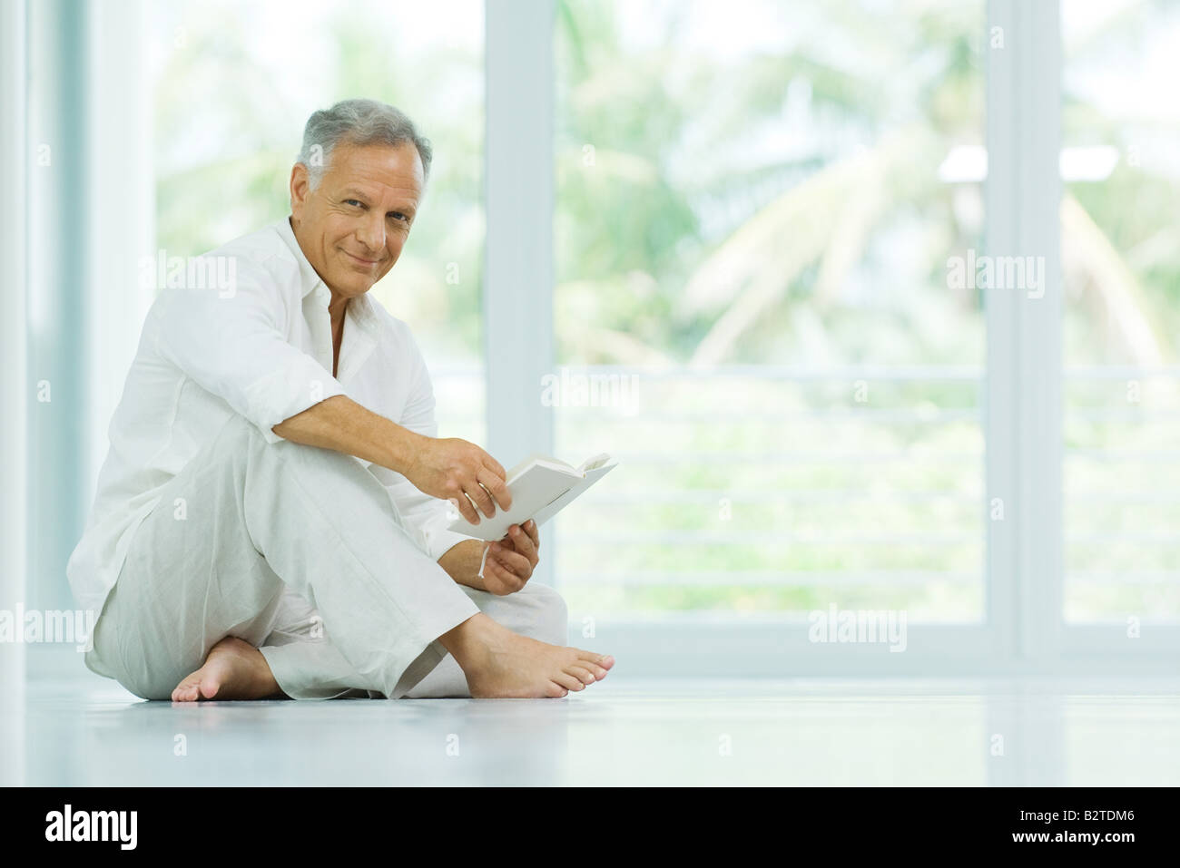 Mature man sitting on the floor, holding book, smiling at camera Stock Photo