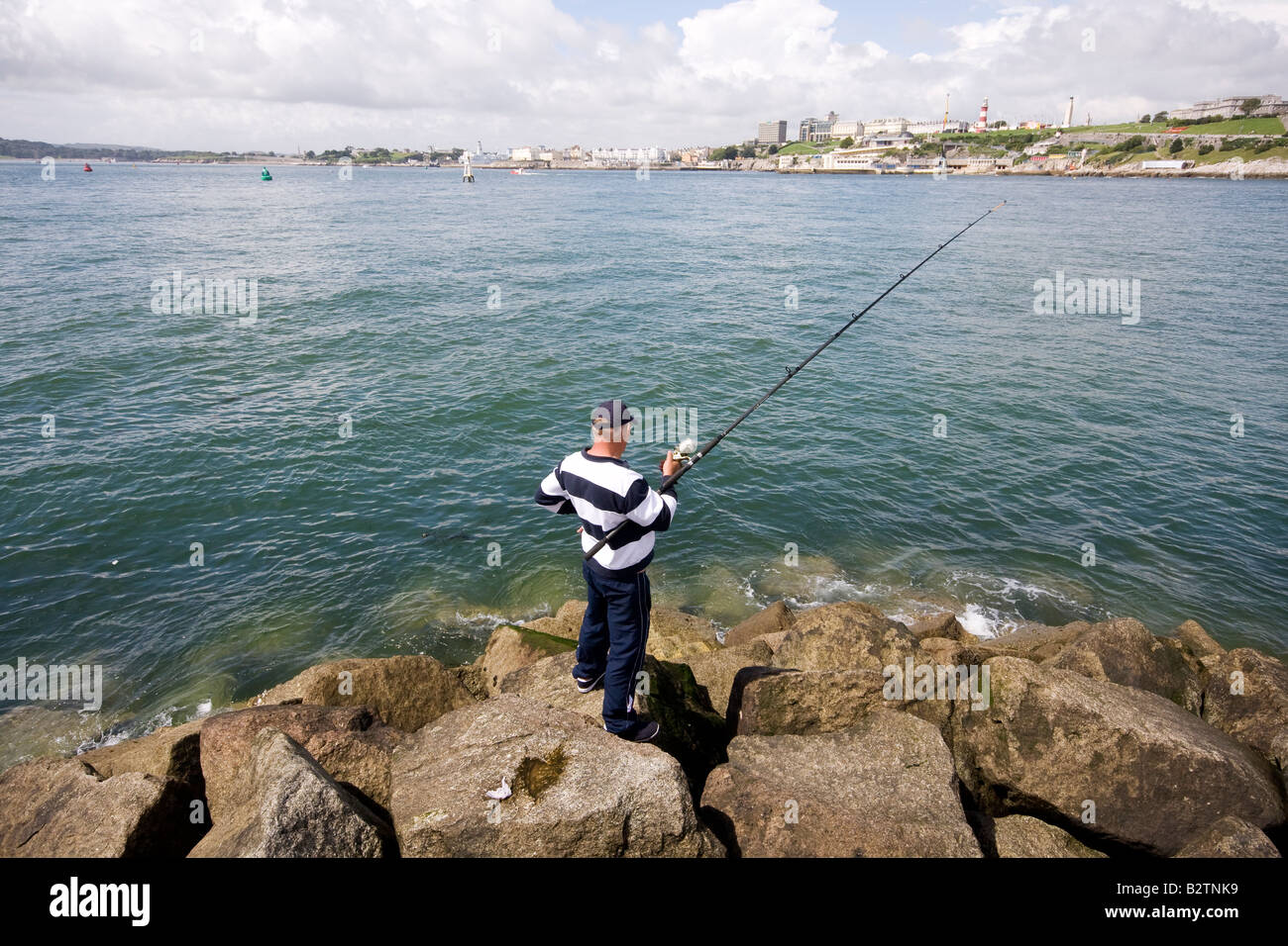 A sea angler fishes from the rocks at the west end of Mountbatten Pier Stock Photo