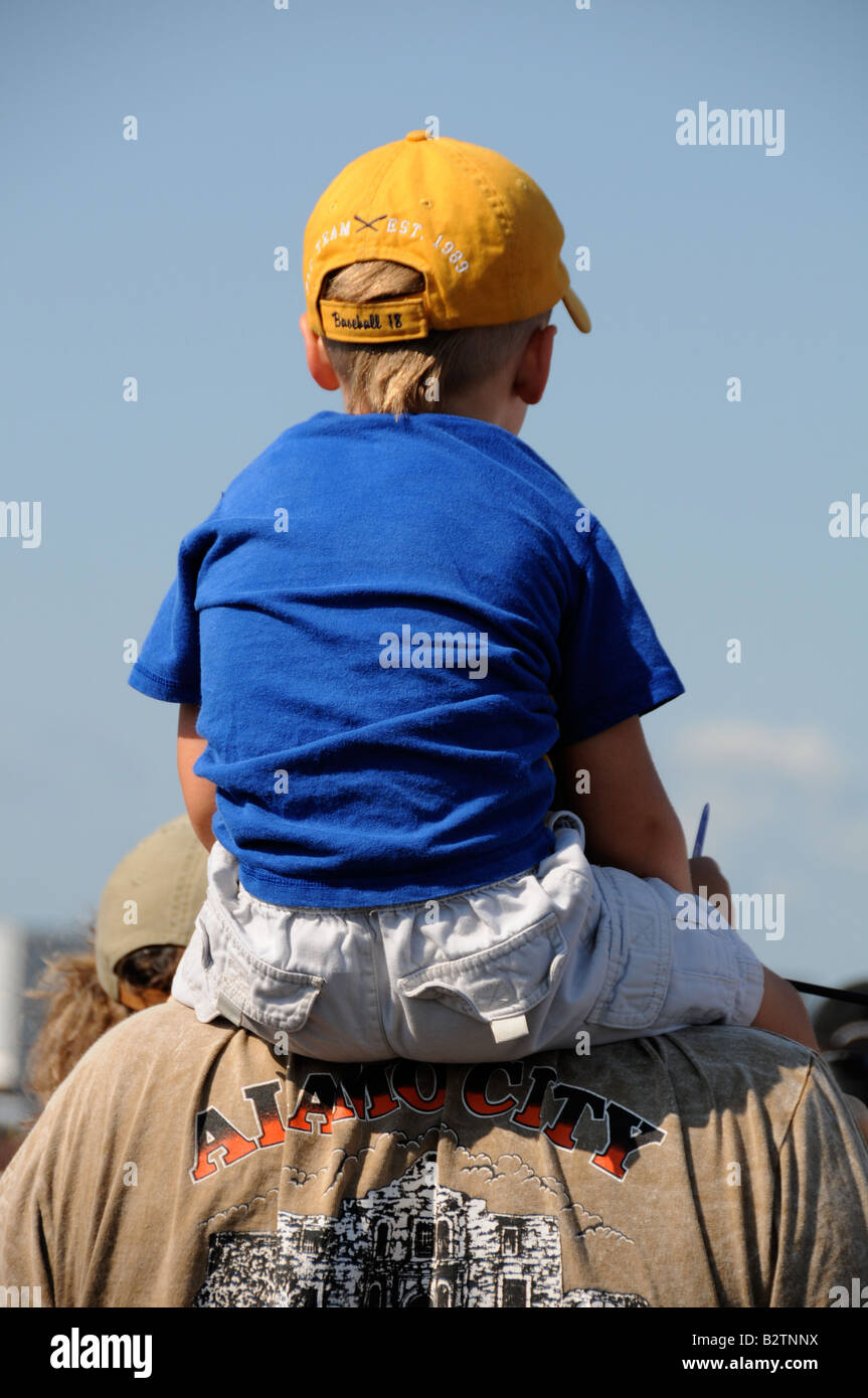 Little boy in yellow cap sitting on fathers shoulder at airshow. Stock Photo