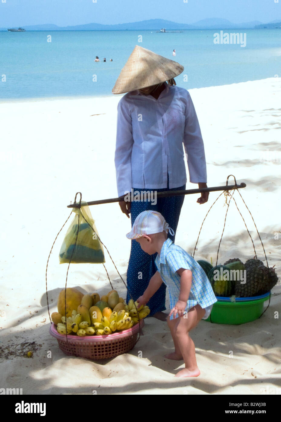 Small boy grabs banana bunch conical hat woman fruit vendor looks on Long Beach Phu Quoc Island Vietnam Stock Photo