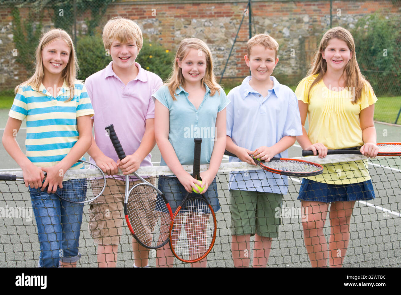 Five young friends with rackets on tennis court smiling Stock Photo