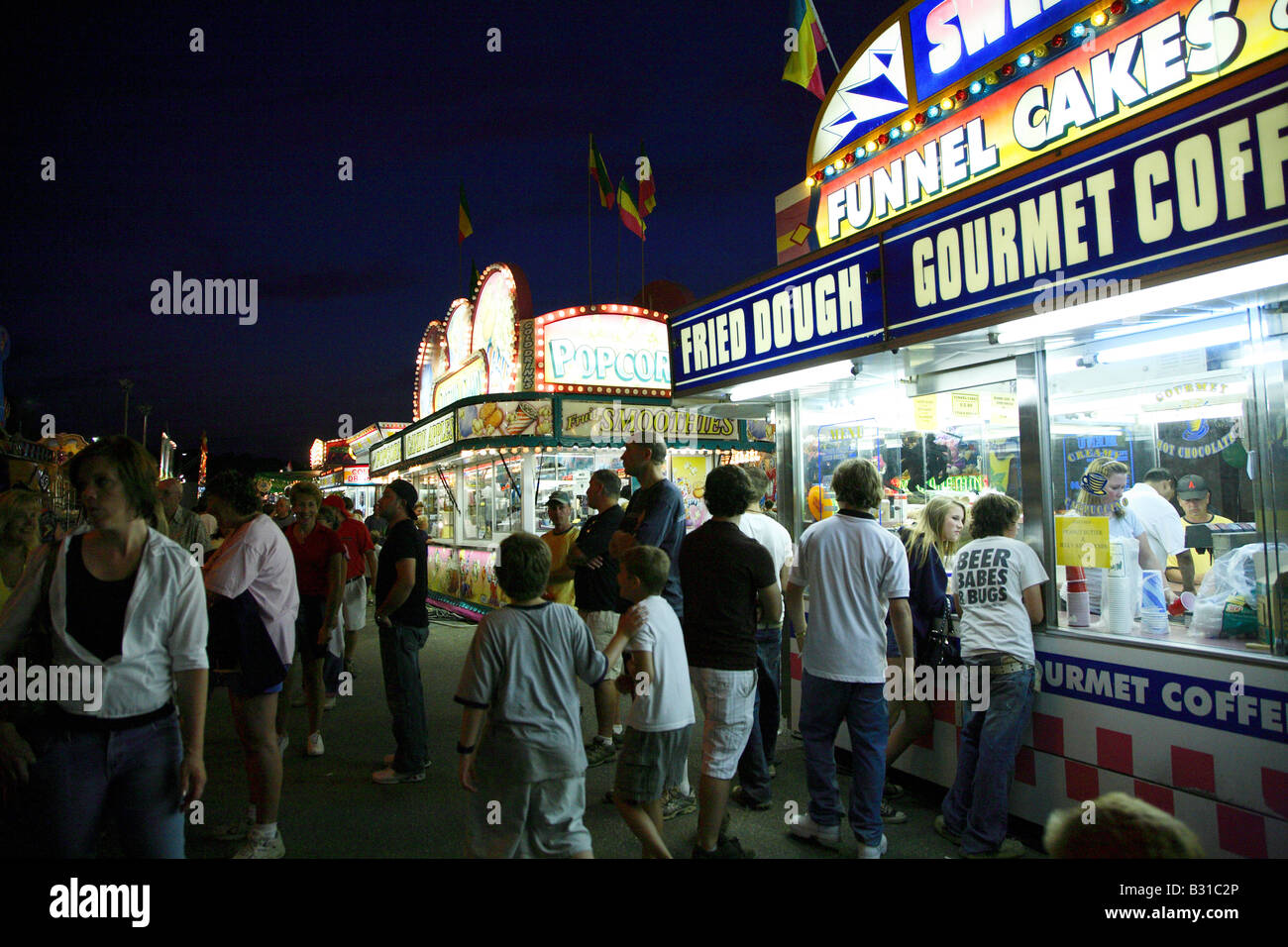 Food stalls at fair lit up at night.  Closest selling funnel cakes and fried dough with popcorn stall behind.  Crowd in dark on Stock Photo