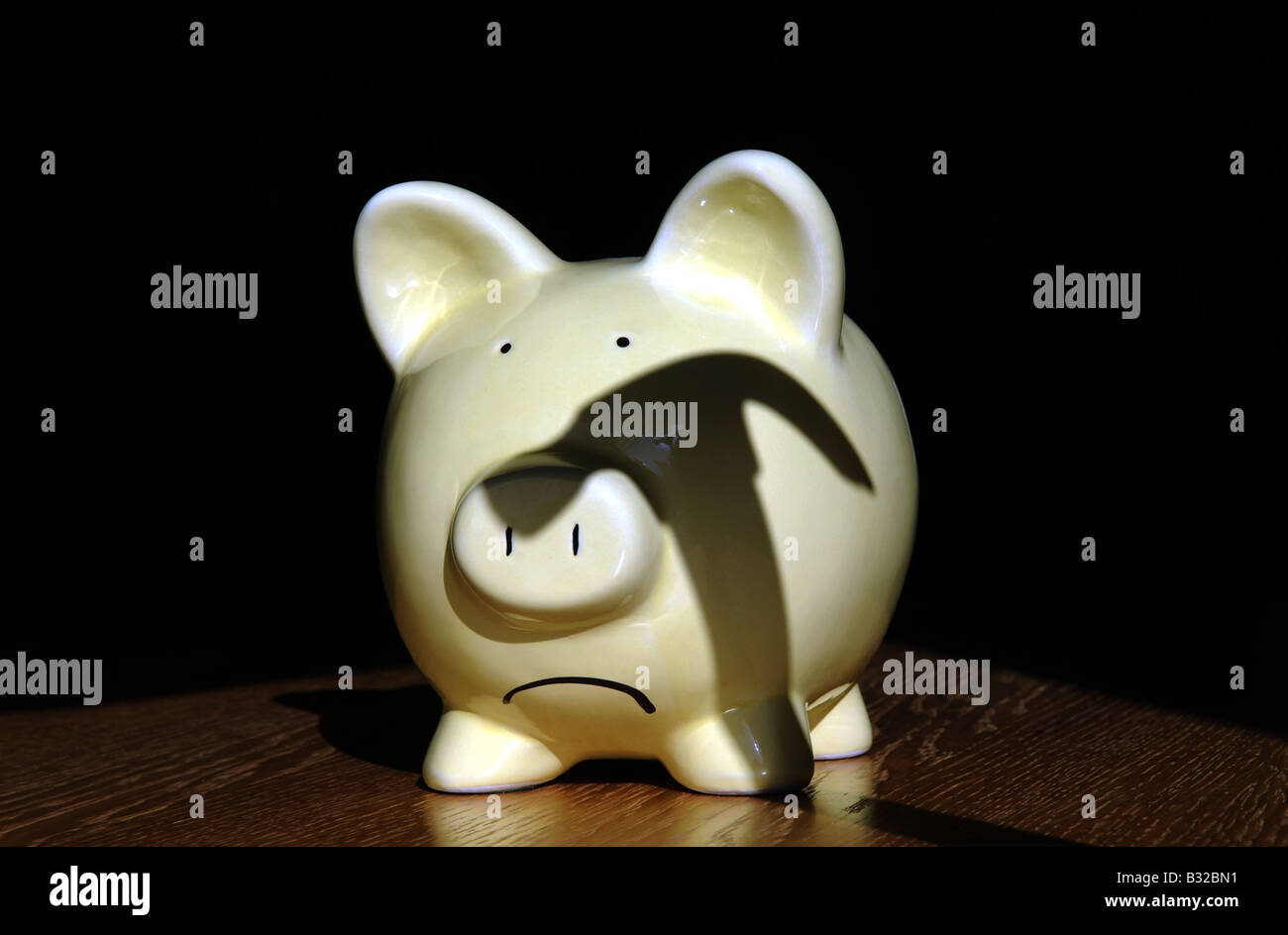 A SAD LOOKING BRITISH PIGGY BANK WITH SHADOW OF A  HAMMER HANGING OVER IT RE THE ECONOMY RECESSION SAVINGS INCOMES WAGES UK. Stock Photo