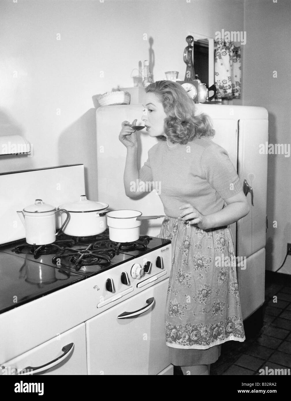 Young woman in an apron in her kitchen tasting her food from a pot Stock Photo