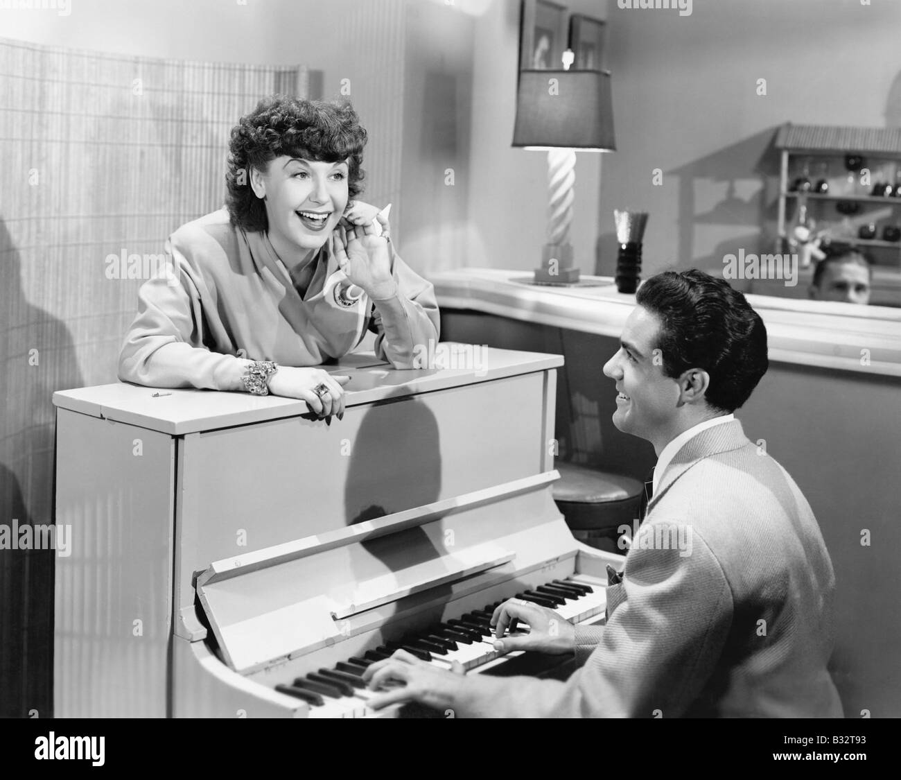 Profile of a young man playing a piano with a young woman standing in front of him and smiling and singing Stock Photo