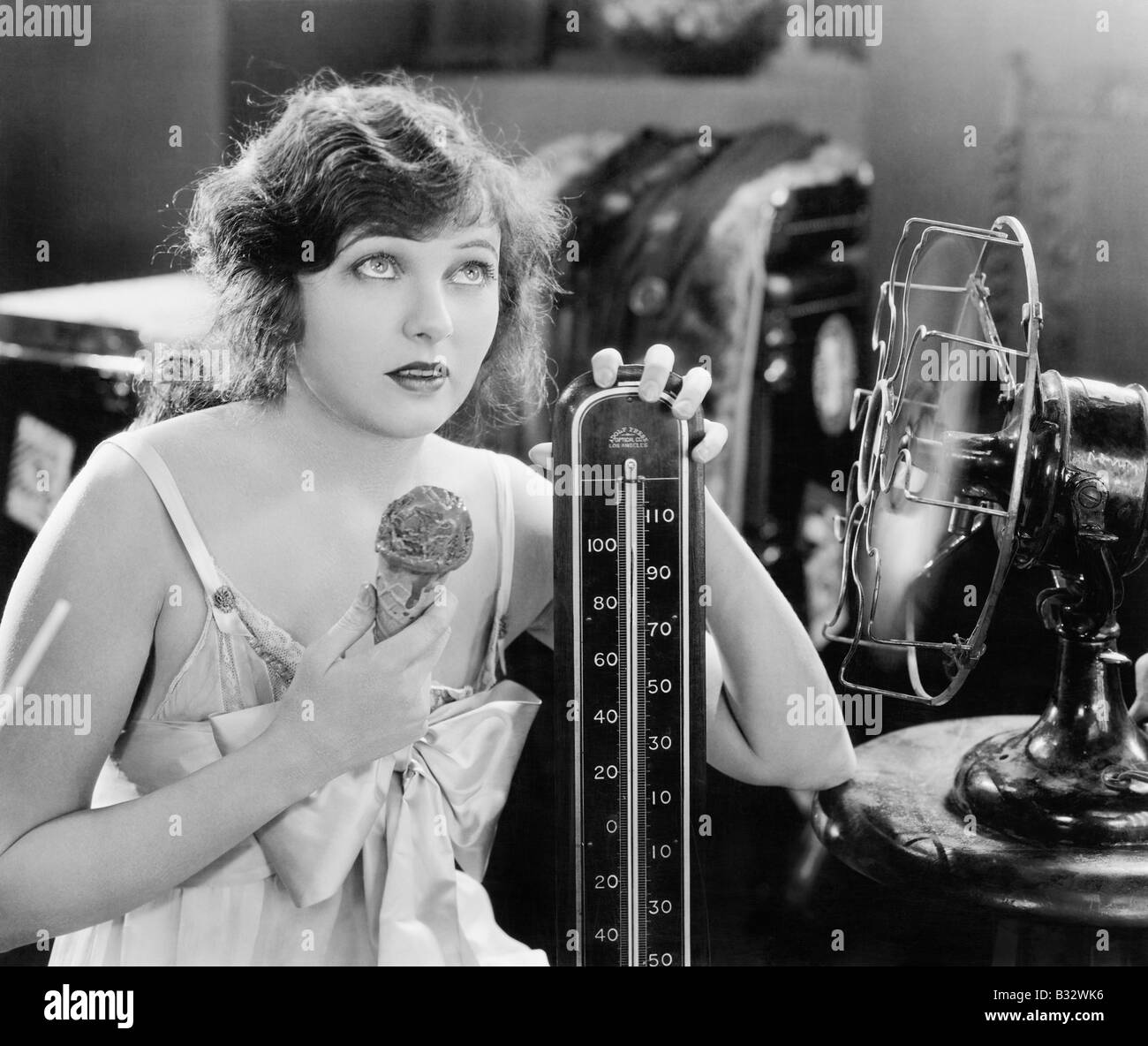 Young woman sitting next to a fan and a thermometer looking hot and eating an ice cream Stock Photo