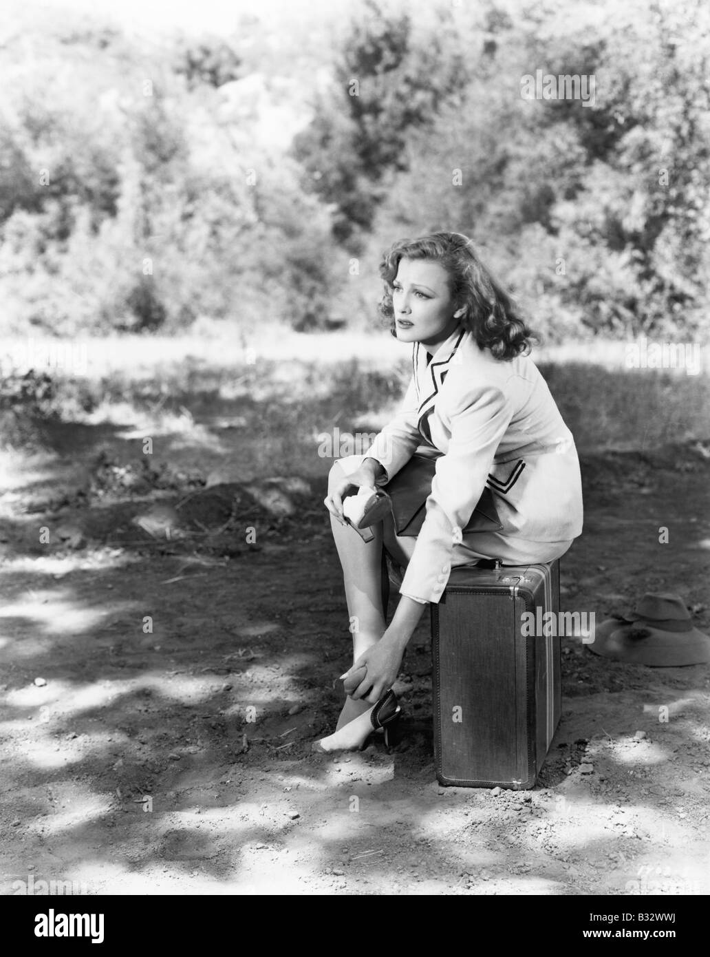 Young woman sitting on the side of the road on her suitcase, rubbing her aching feet Stock Photo