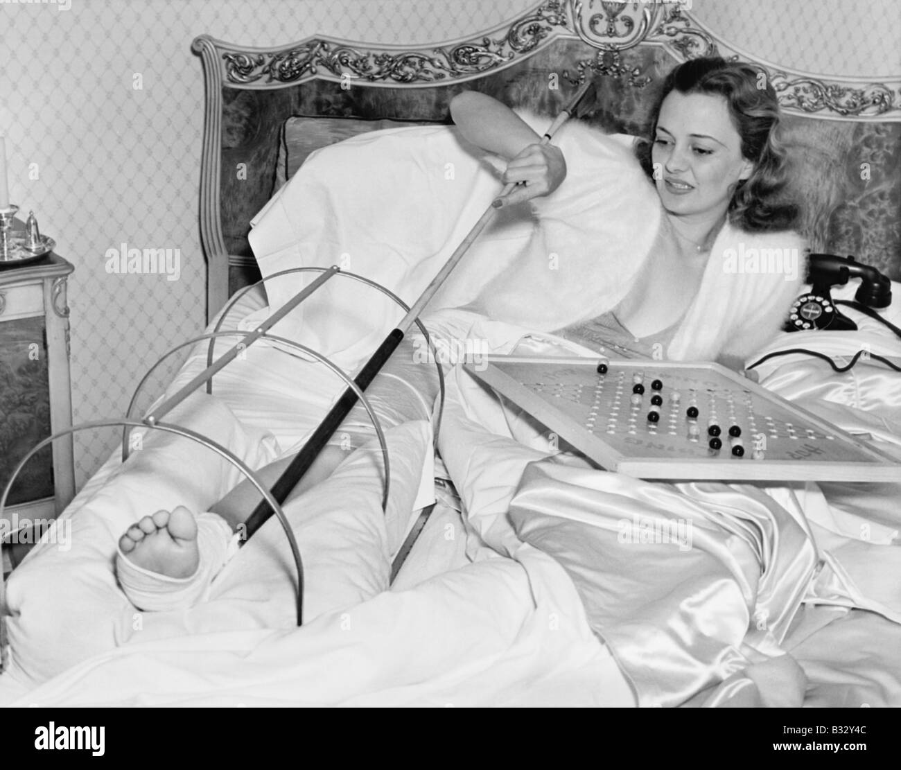 Woman in bed trying to scratch her broken foot with a pole Stock Photo