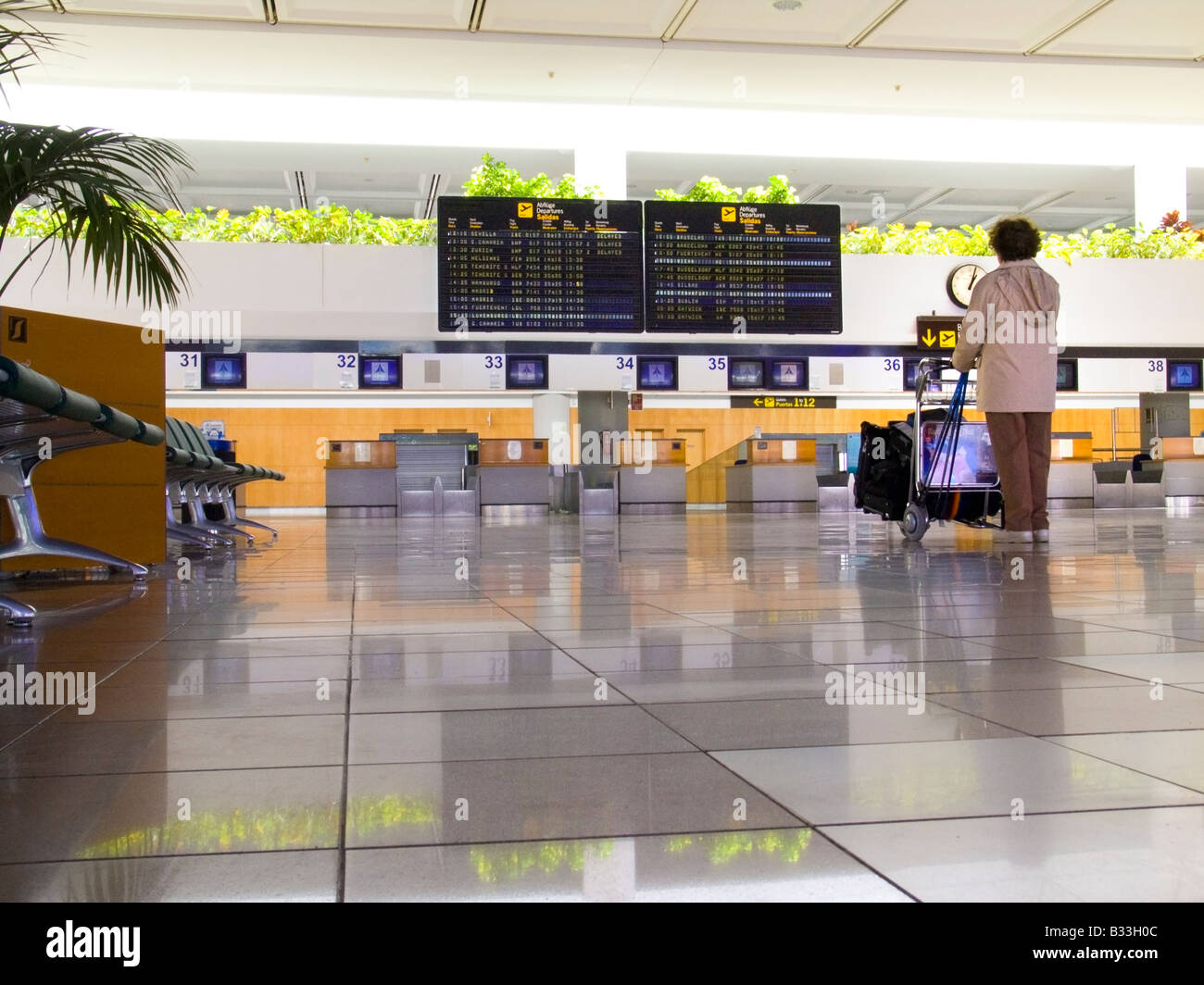 Airline passenger with luggage on trolley standing alone waiting on deserted airport concourse looking at information boards Stock Photo