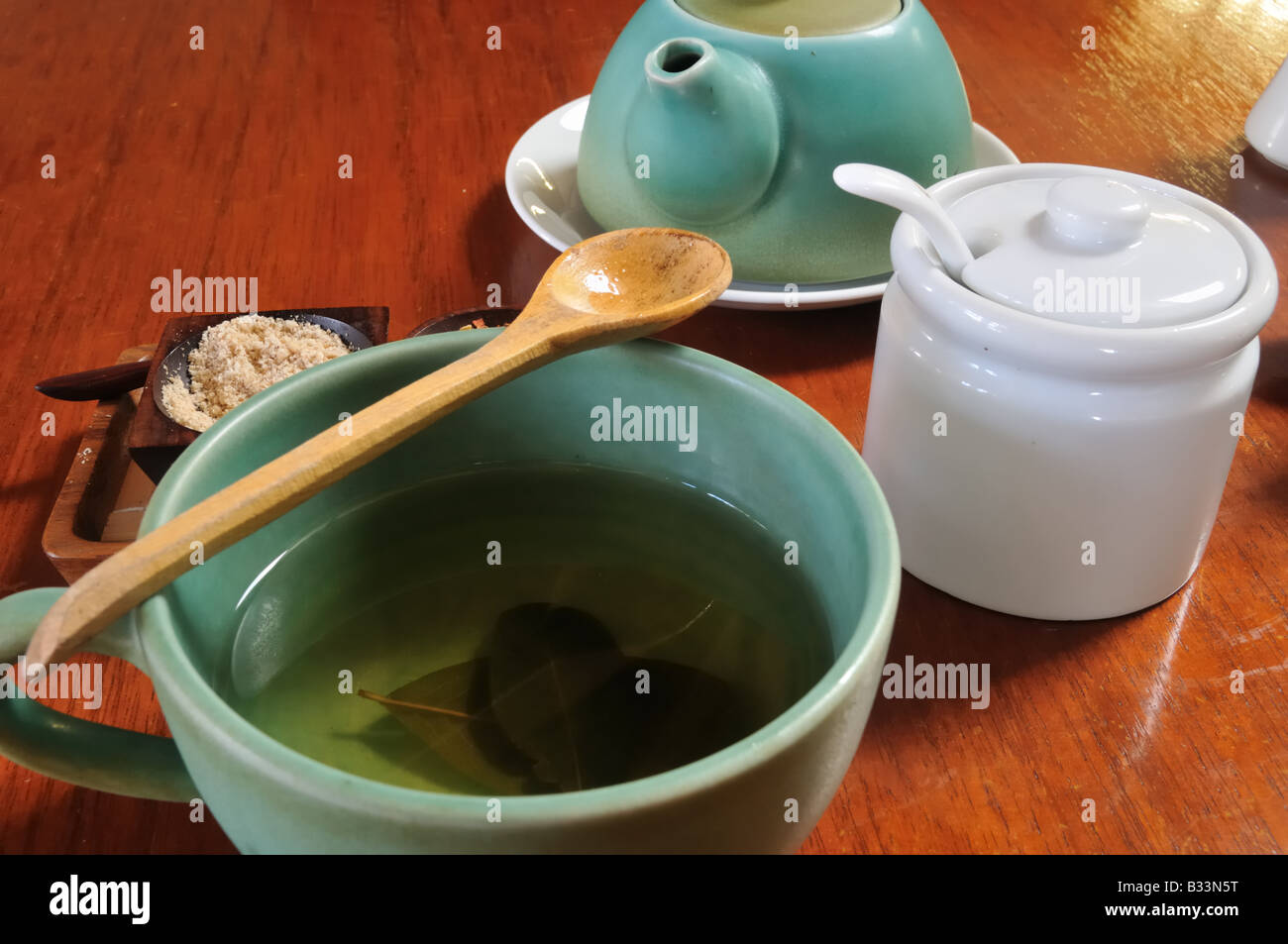 A coca tea set with a sugar dish, kettle and wooden spoon sit on a rich wood table.  Coca tea leaves are visible steeping. Stock Photo