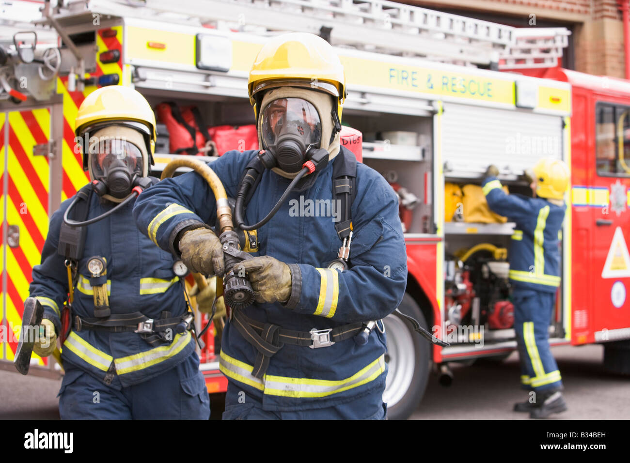 Two firefighters with hose and axe walking away from fire engine and another firefighter in background (selective focus) Stock Photo