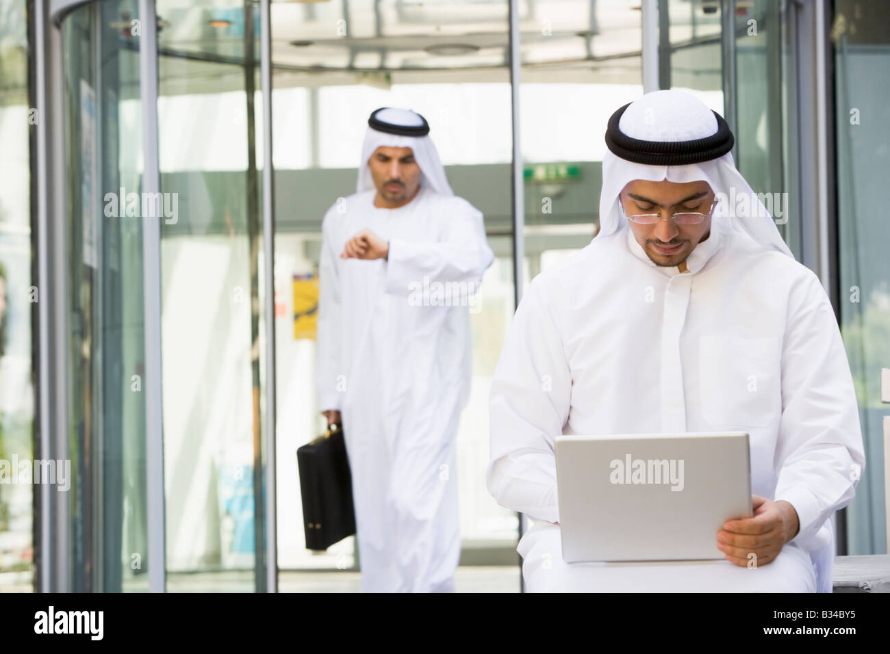 Businessman outdoors using laptop with businessman walking in background checking watch (selective focus) Stock Photo