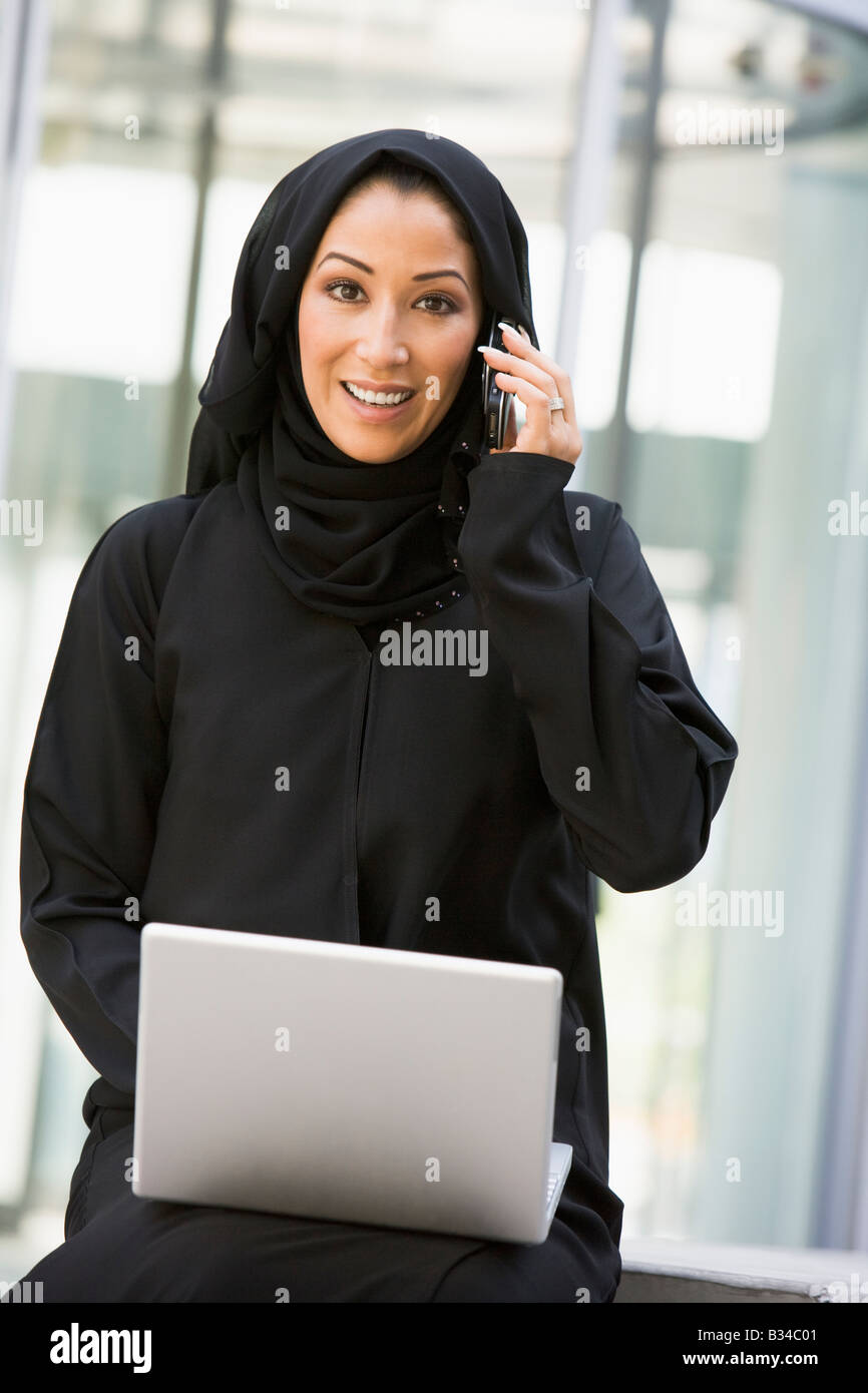 Businesswoman sitting outdoors by building with laptop using cellular phone smiling (selective focus) Stock Photo