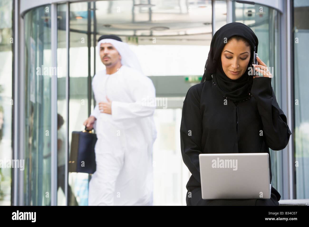 Businesswoman sitting outdoors by building with laptop using cellular phone with businessman running in background Stock Photo