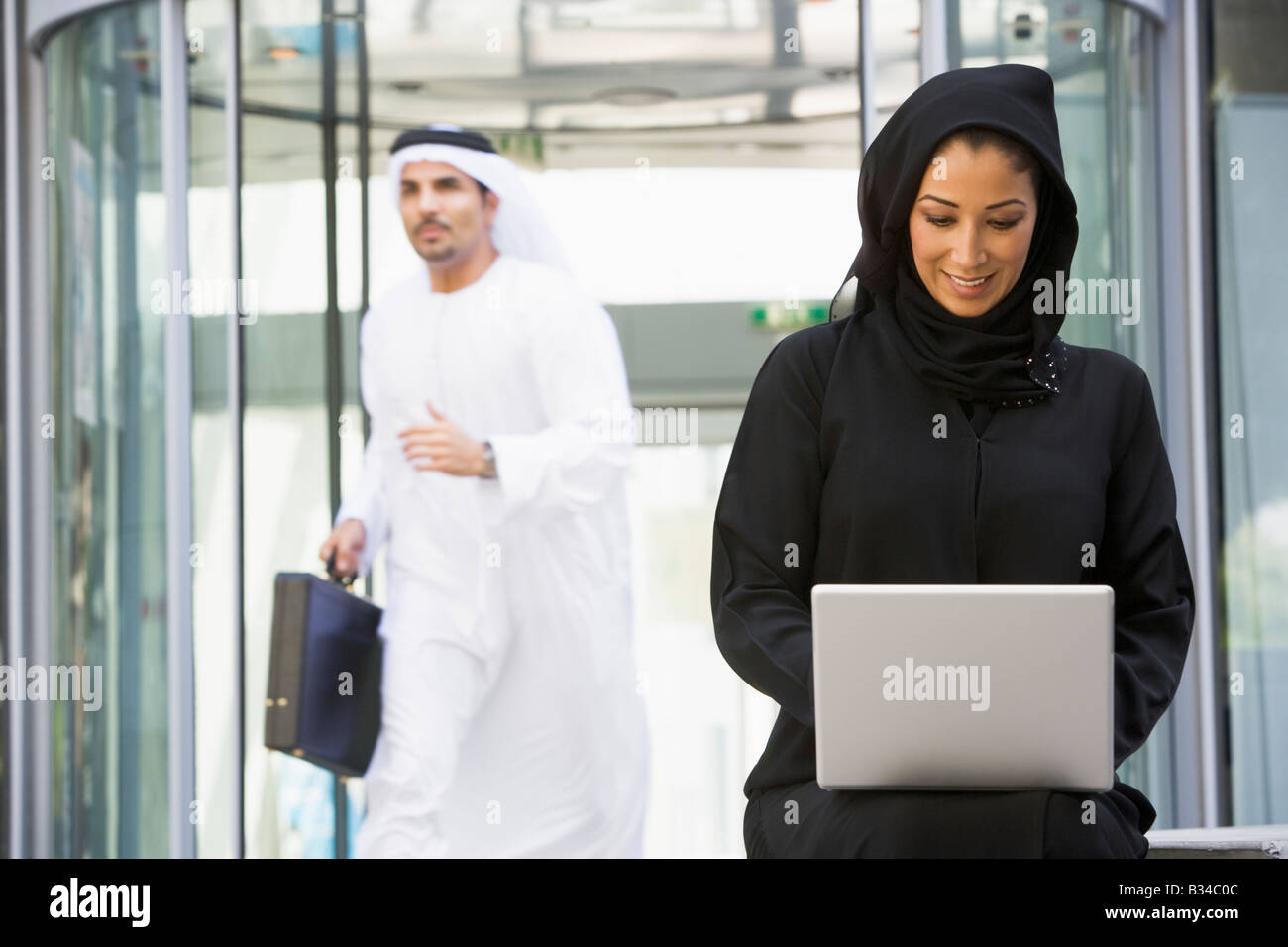 Businesswoman sitting outdoors by building with laptop smiling with businessman running in background (selective focus) Stock Photo