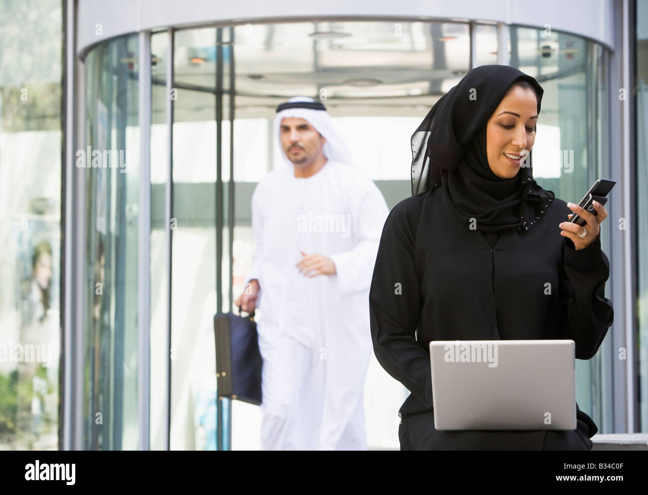 Businesswoman sitting outdoors by building with laptop holding cellular phone with businessman running in background Stock Photo