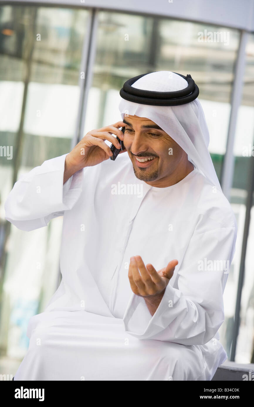 Businessman sitting outdoors by building using cellular phone and smiling (selective focus) Stock Photo