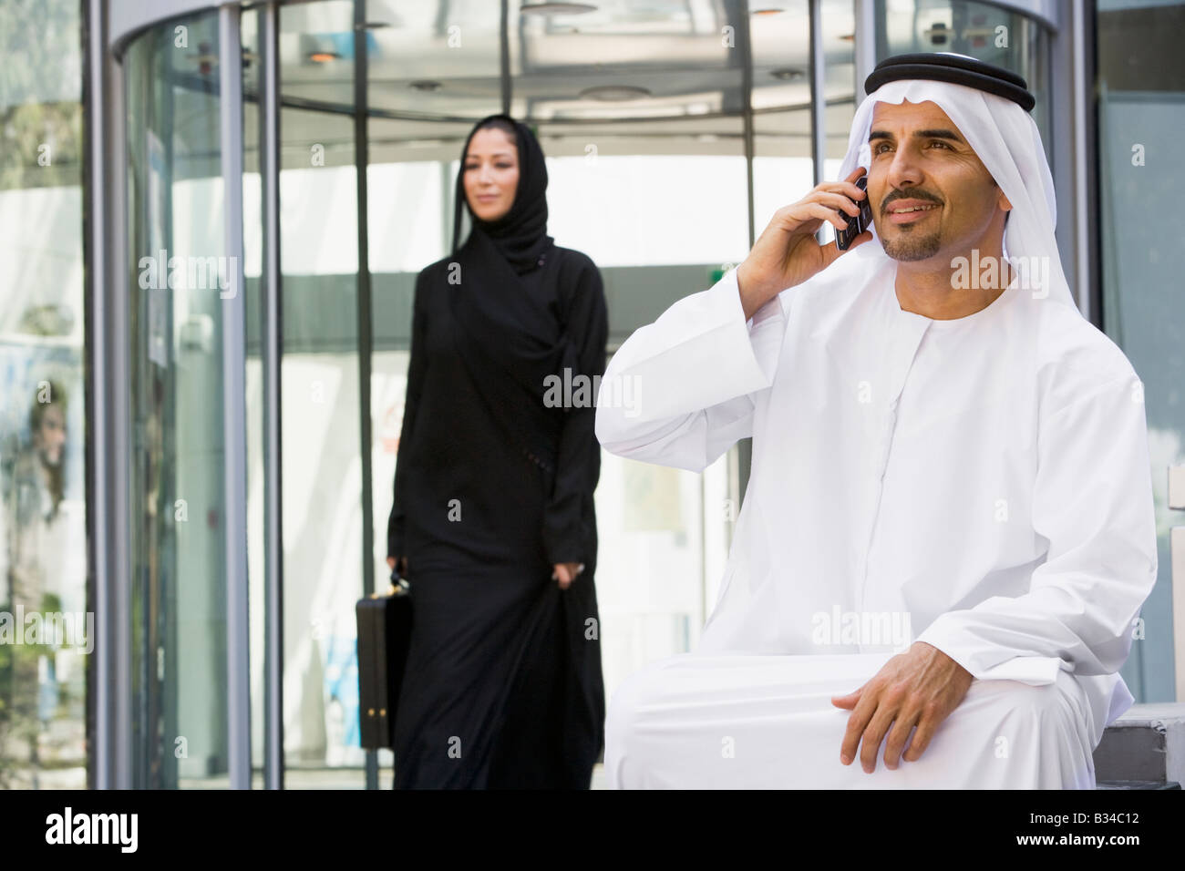 Businessman sitting outdoors by building using cellular phone with businesswoman walking in background (selective focus) Stock Photo