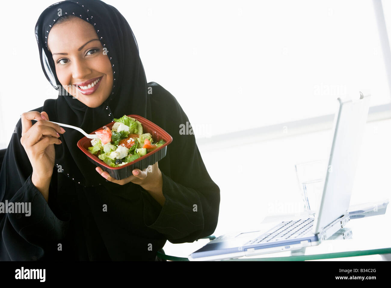 Woman eating salad near laptop smiling (high key/selective focus) Stock Photo