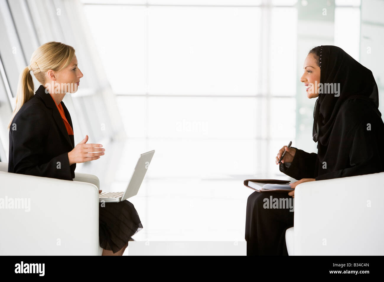 Two businesswomen indoors with a laptop talking and smiling (high key/selective focus) Stock Photo