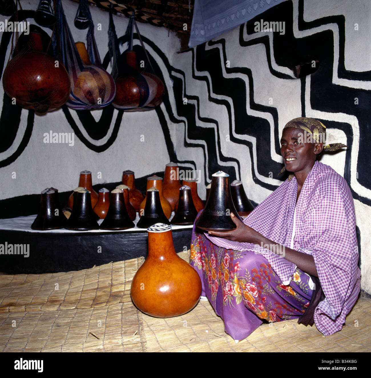 Uganda, Southern Uganda, Mbarara. A Bahima woman with her family's wooden milk pots (known as ekyanzi) and gourds, which are kept on a special raised step in a corner of the living room. Houses are often decorated with original African designs.The Bahima are Banyarwanda-speaking people live near the Rwanda border in southern Uganda. They keep long-horned Ankole cattle, an African taurine breed with origins dating back prior to the introduction of humped-back or zebu cattle into the Horn of Africa during the human invasions from Arabia in the seventh century BC. Stock Photo