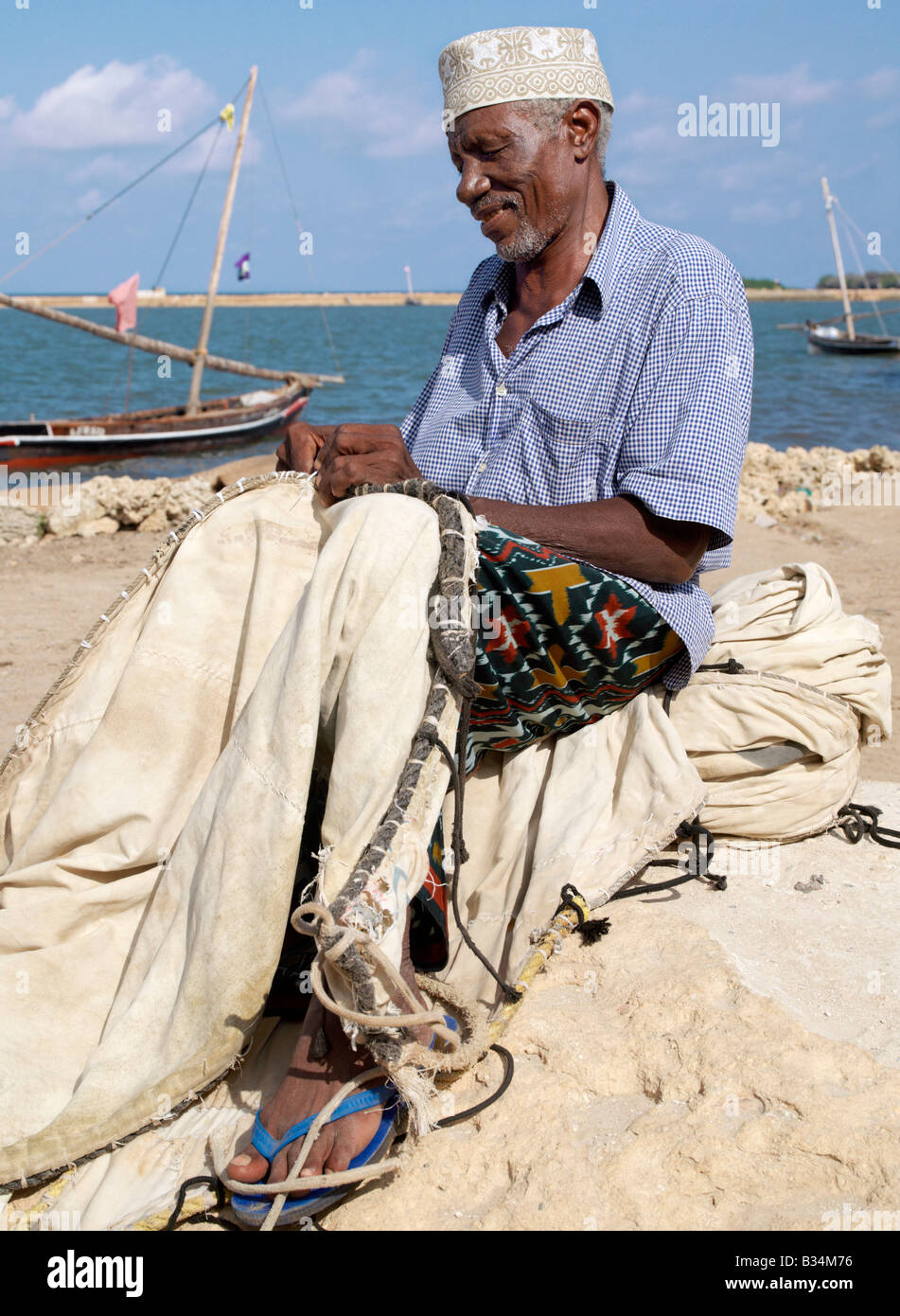 Kenya, Kisingitini, Pate Island. A fisherman repairs the sail of his wooden sailing boat, known as mashua, along the waterfront Stock Photo